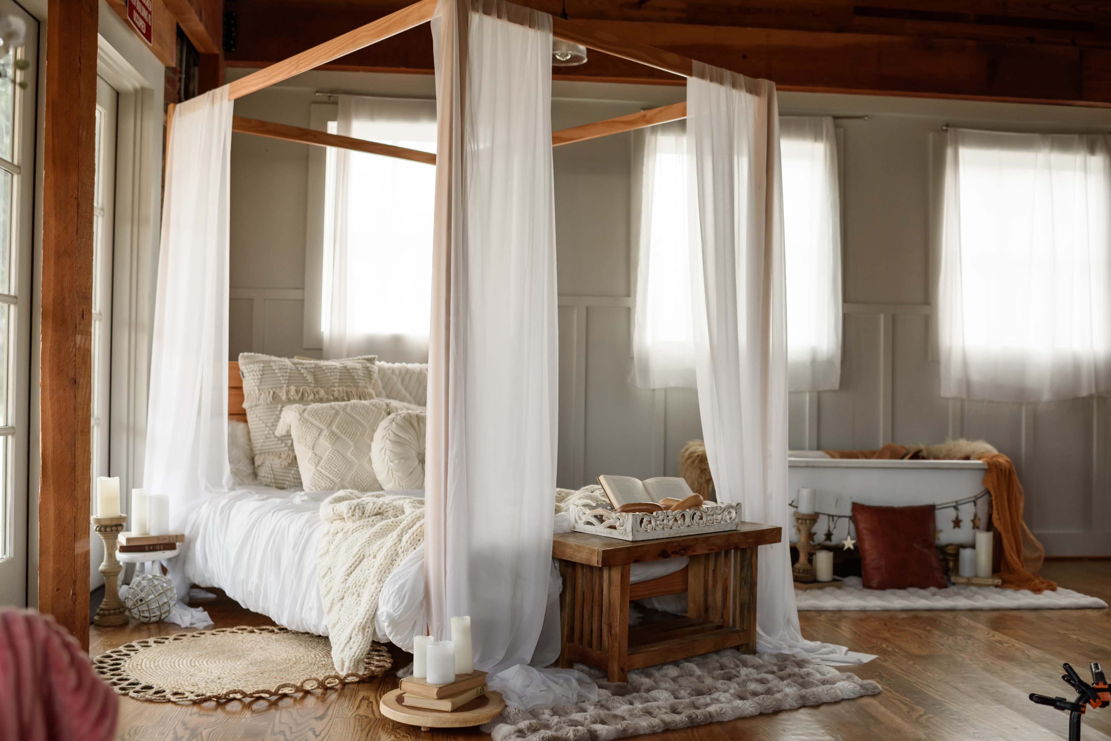The image shows a cozy bedroom with a canopy bed draped in white fabric, surrounded by candles, a wooden side table, and a vintage bathtub in the background.