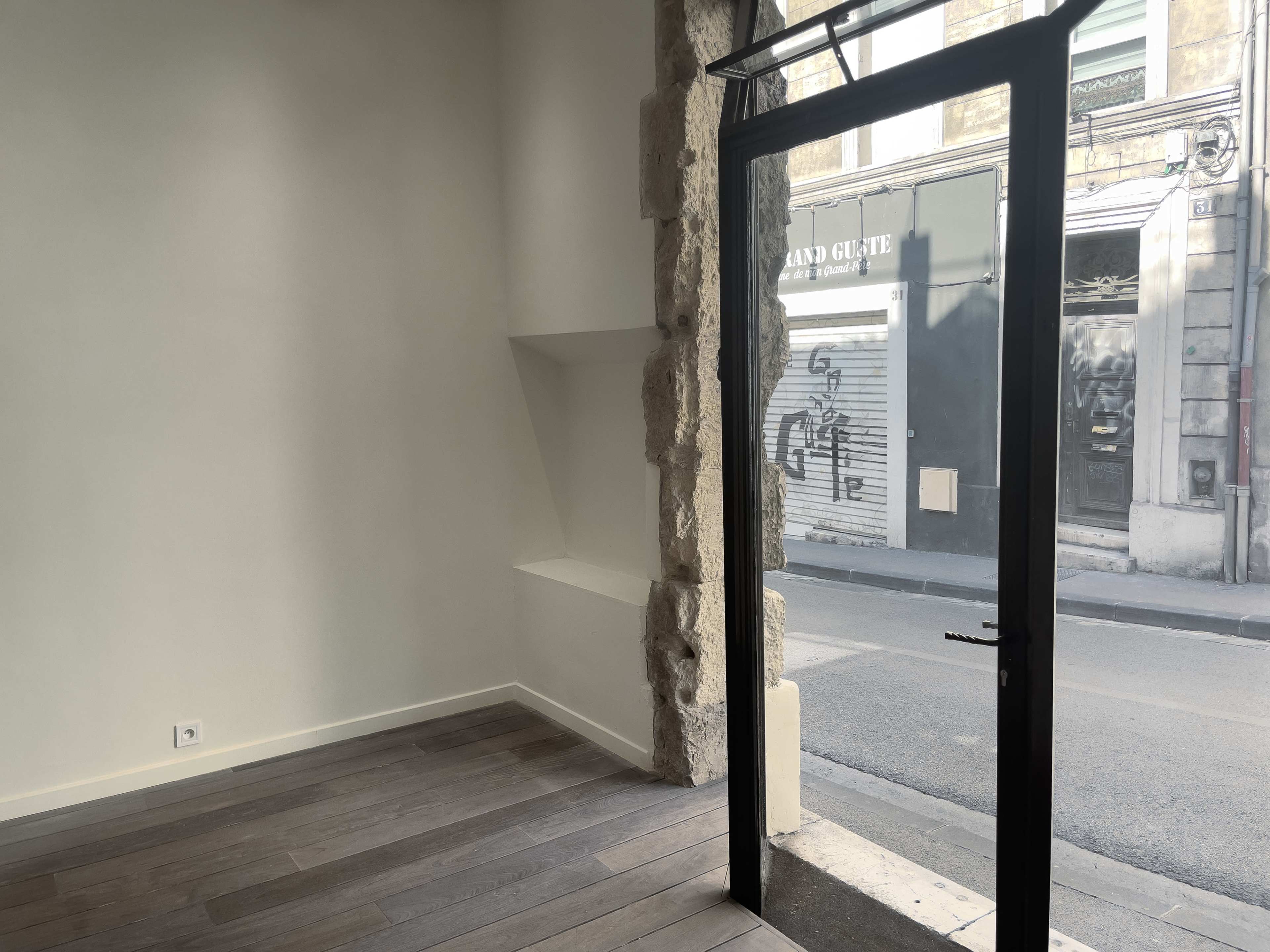 The image shows the interior of a vacant retail space with large windows and a stone wall, looking out onto a street.