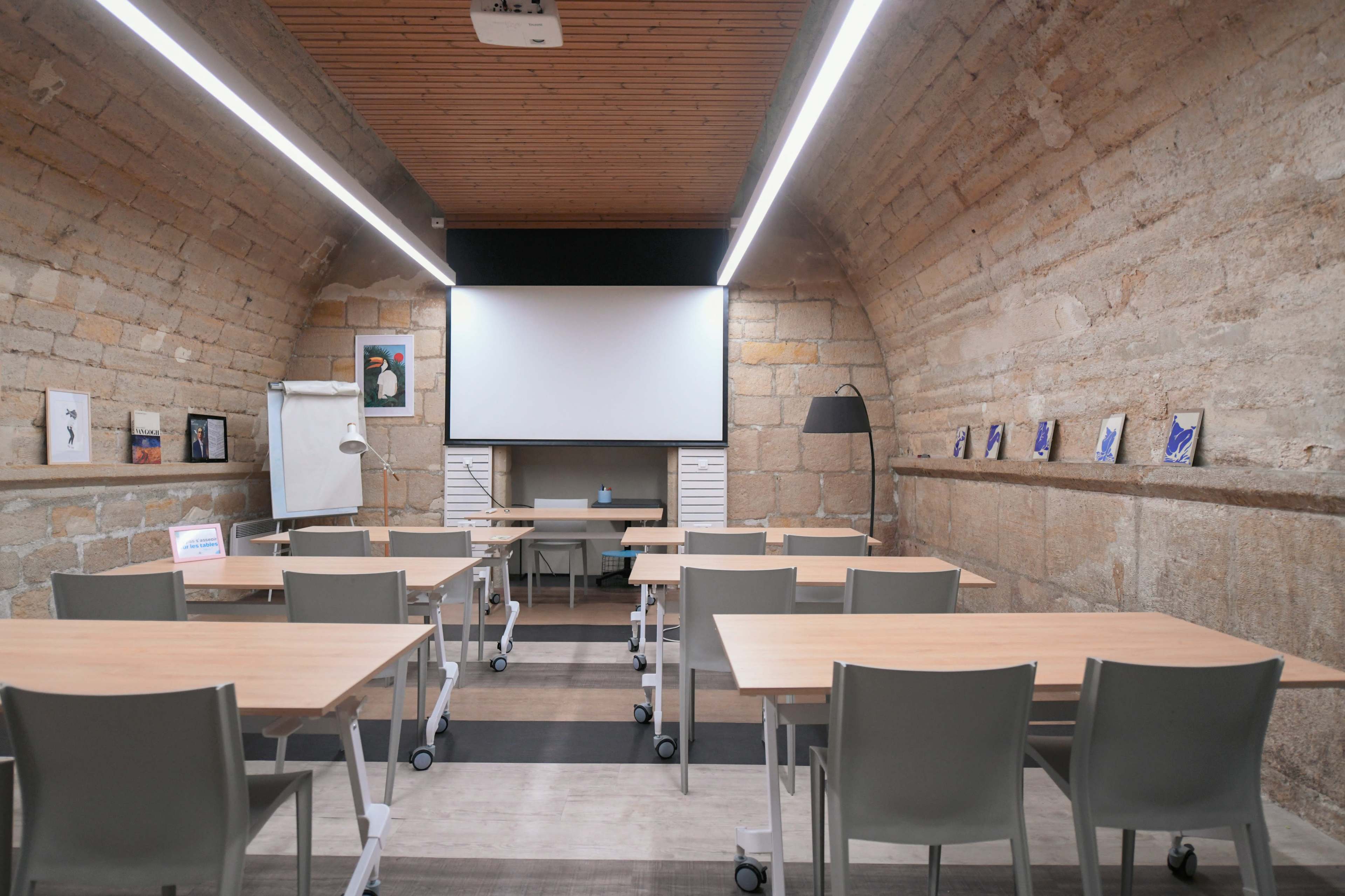 A classroom with wooden tables and chairs arranged in front of a blank projector screen, set against a stone wall with framed artworks.