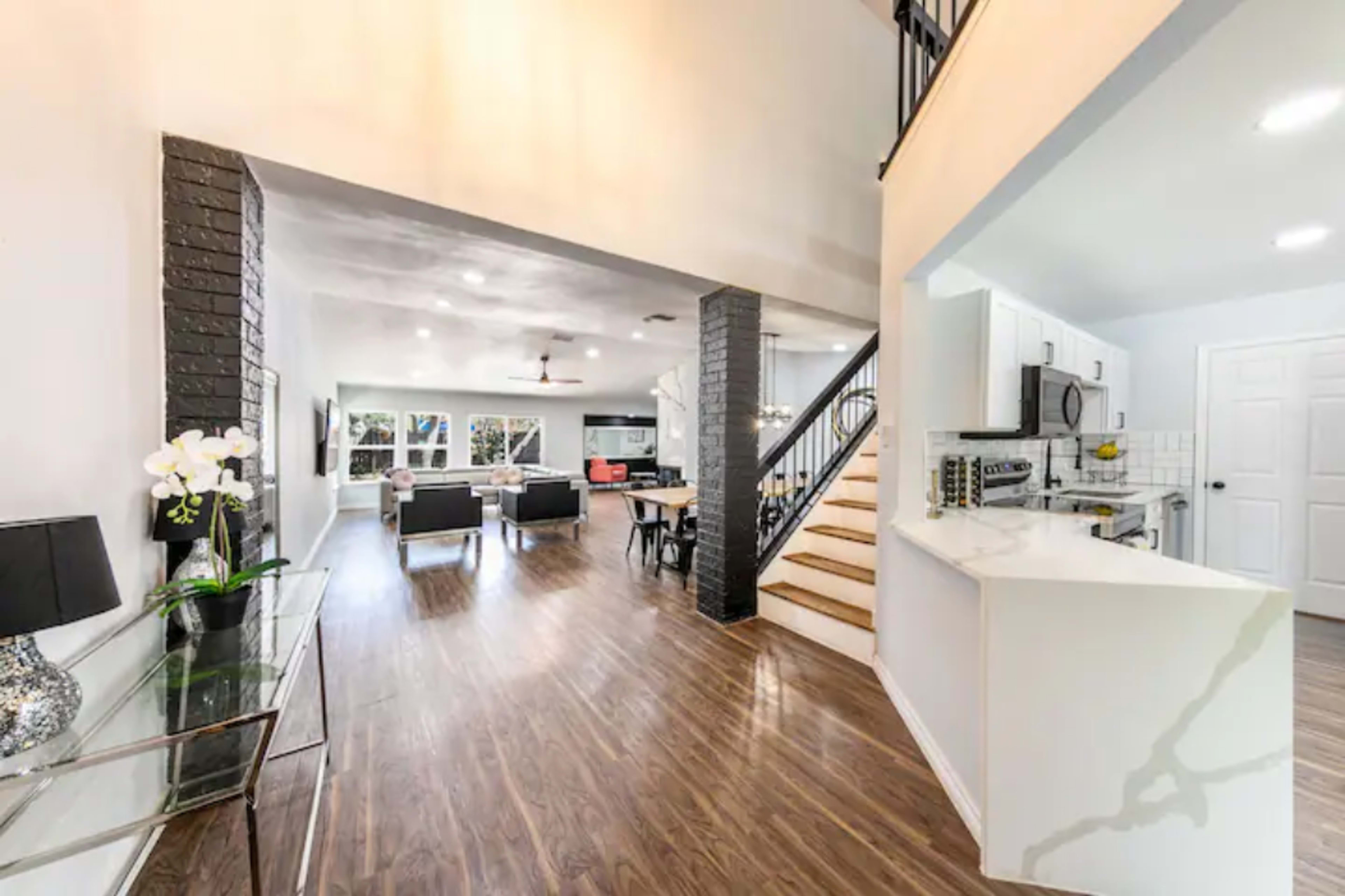 A modern interior space with an open layout, featuring a staircase on the right, a kitchen area, and a living area with seating and a dining table.