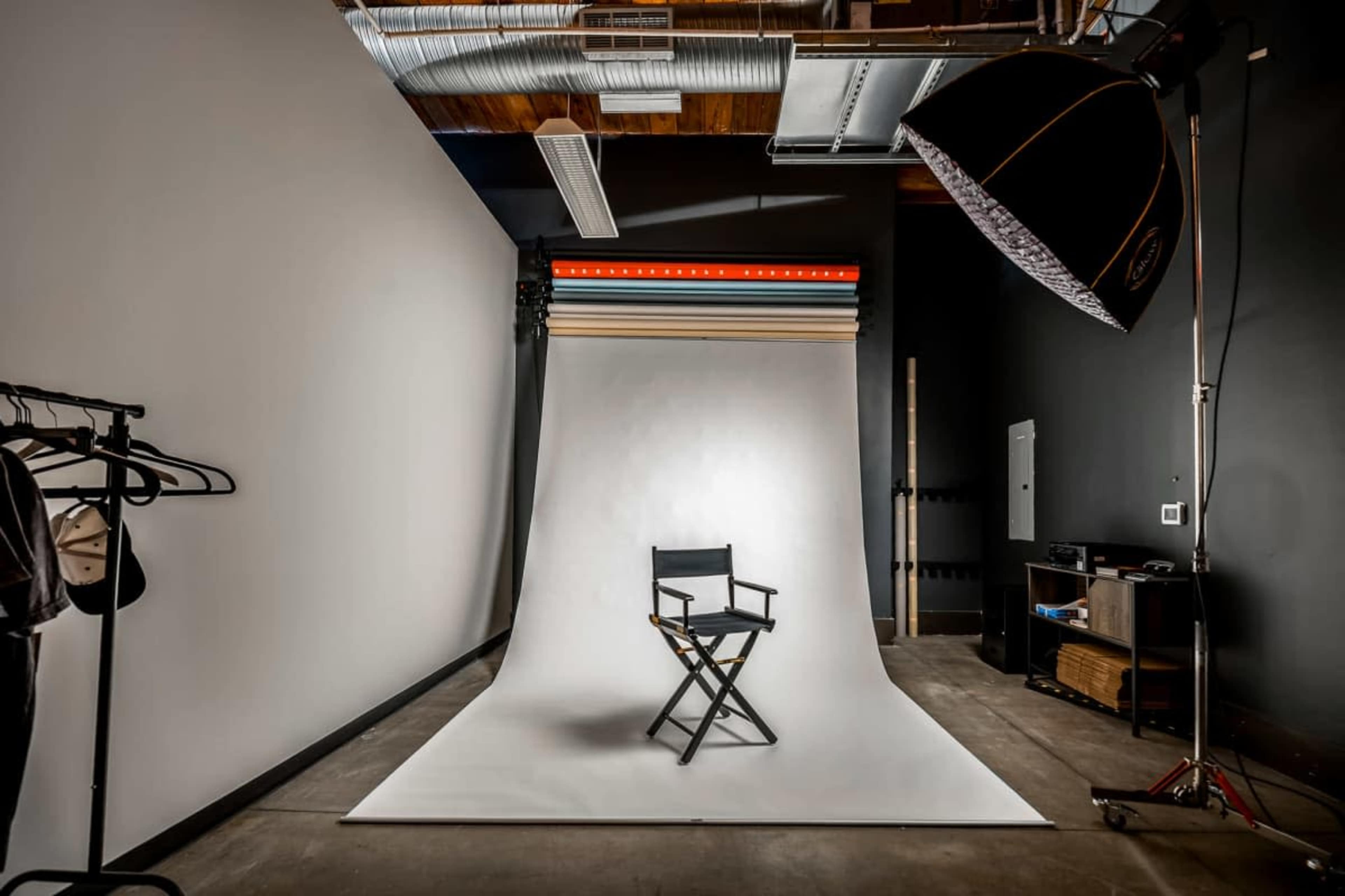 The image shows a photography studio with a director's chair in front of a white backdrop, illuminated by soft lighting.