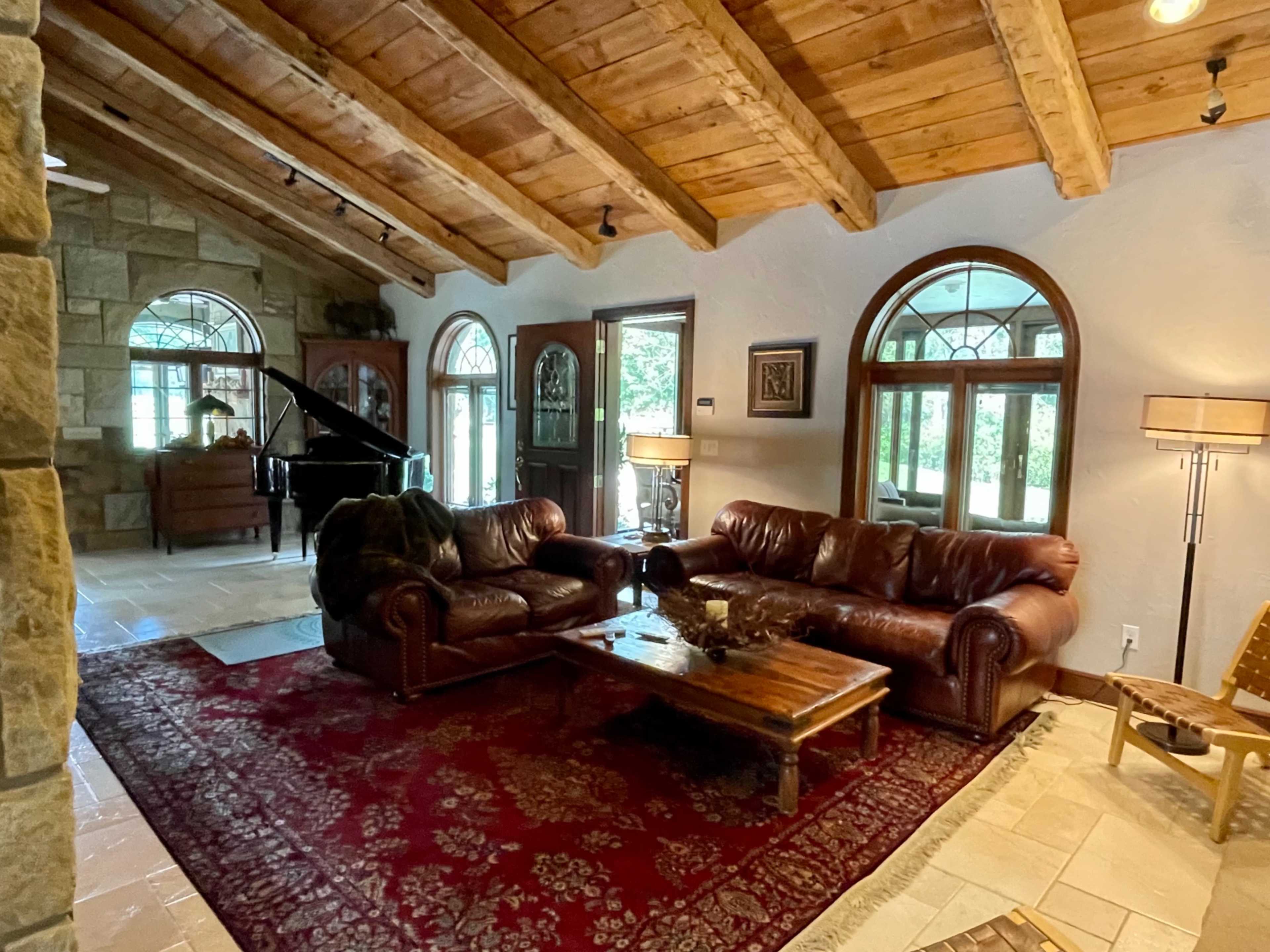 A rustic living room with wooden beams, leather sofas, a coffee table, and a grand piano visible in the background through arched windows.