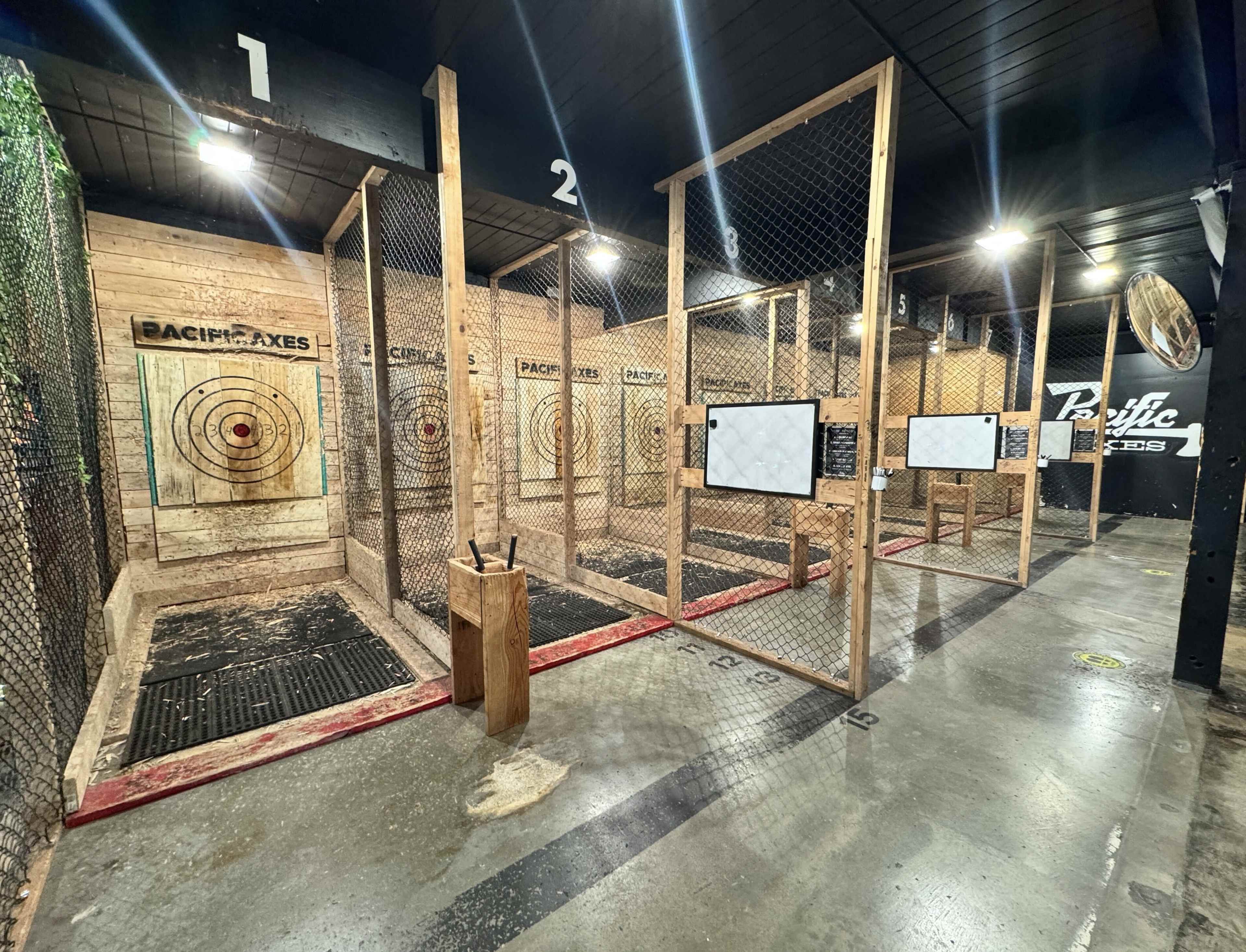 The image shows an indoor axe-throwing venue with several lanes, each enclosed by wooden barriers, featuring target boards and a concrete floor.