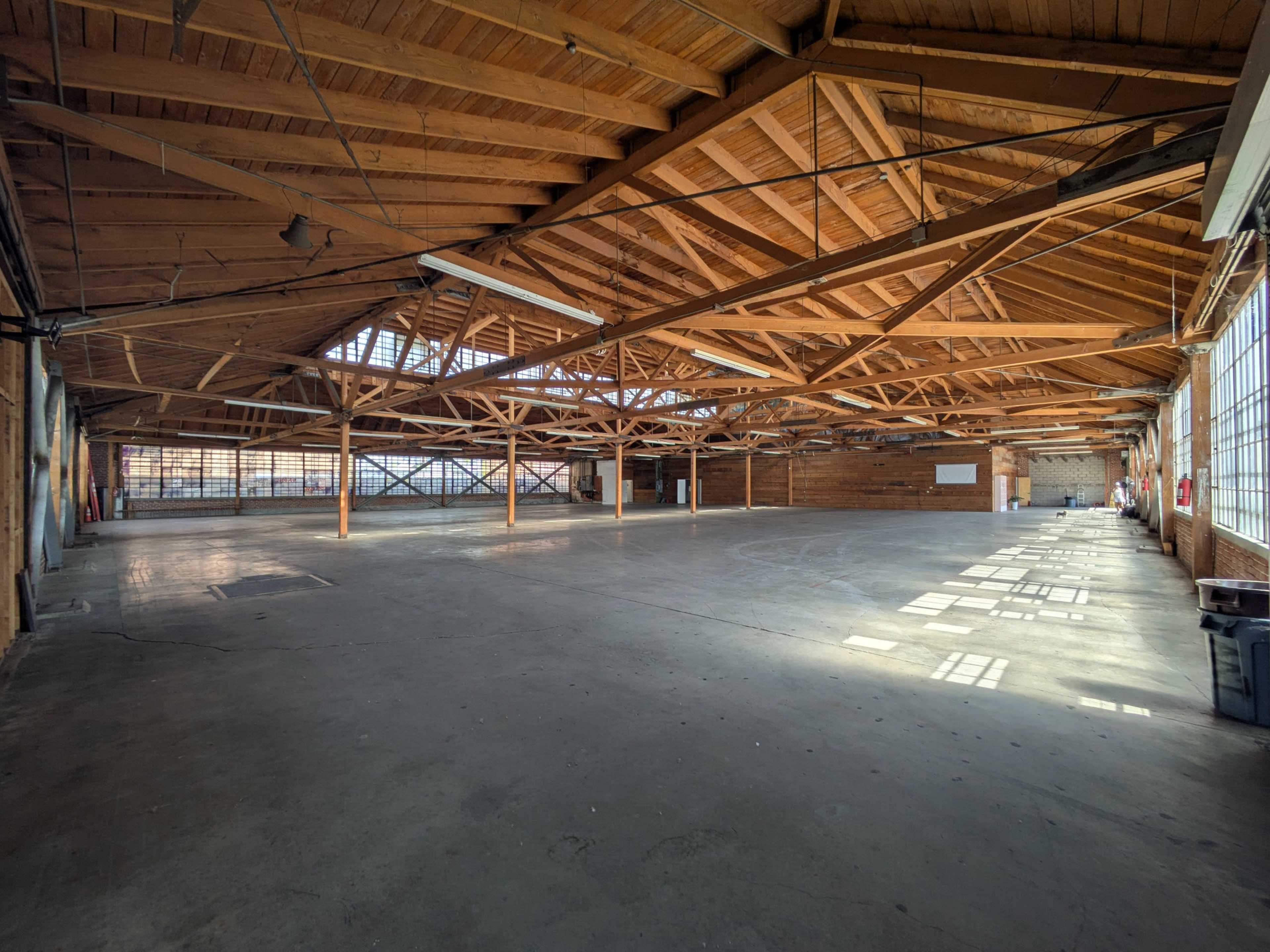 The image shows a large, empty warehouse with a wooden beamed ceiling and concrete flooring, illuminated by natural light from large windows.