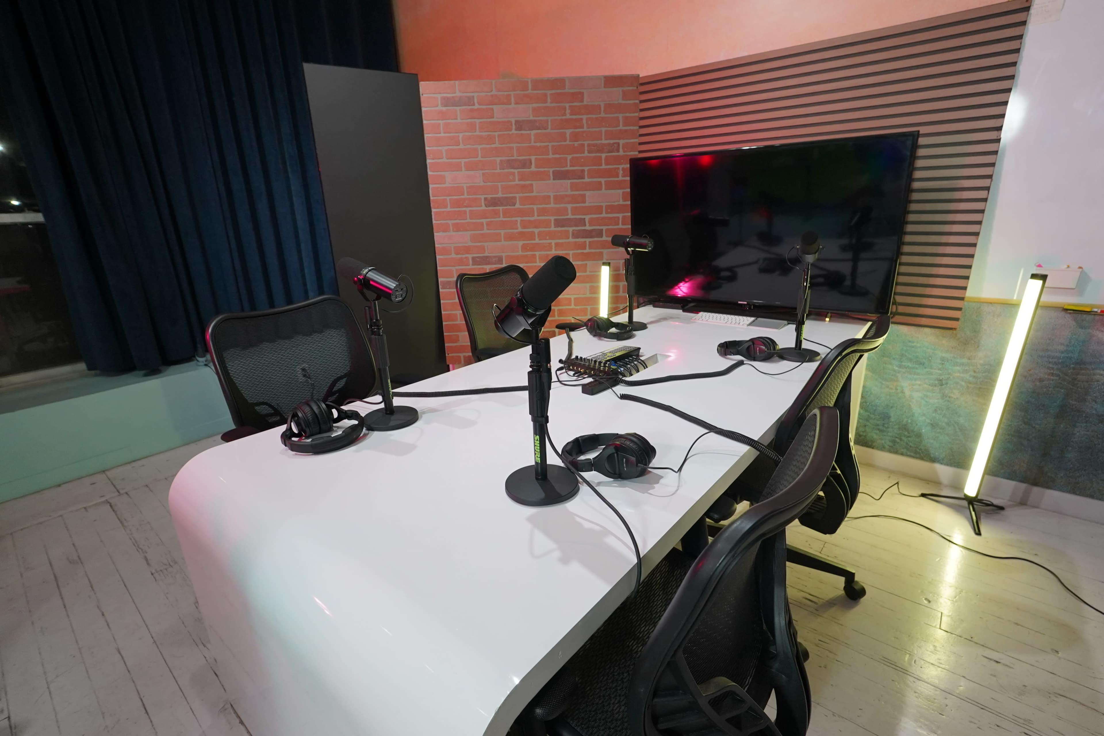 A modern podcast studio features a white table with multiple microphones and headphones, set against a colorful backdrop with a large screen and brick wall.