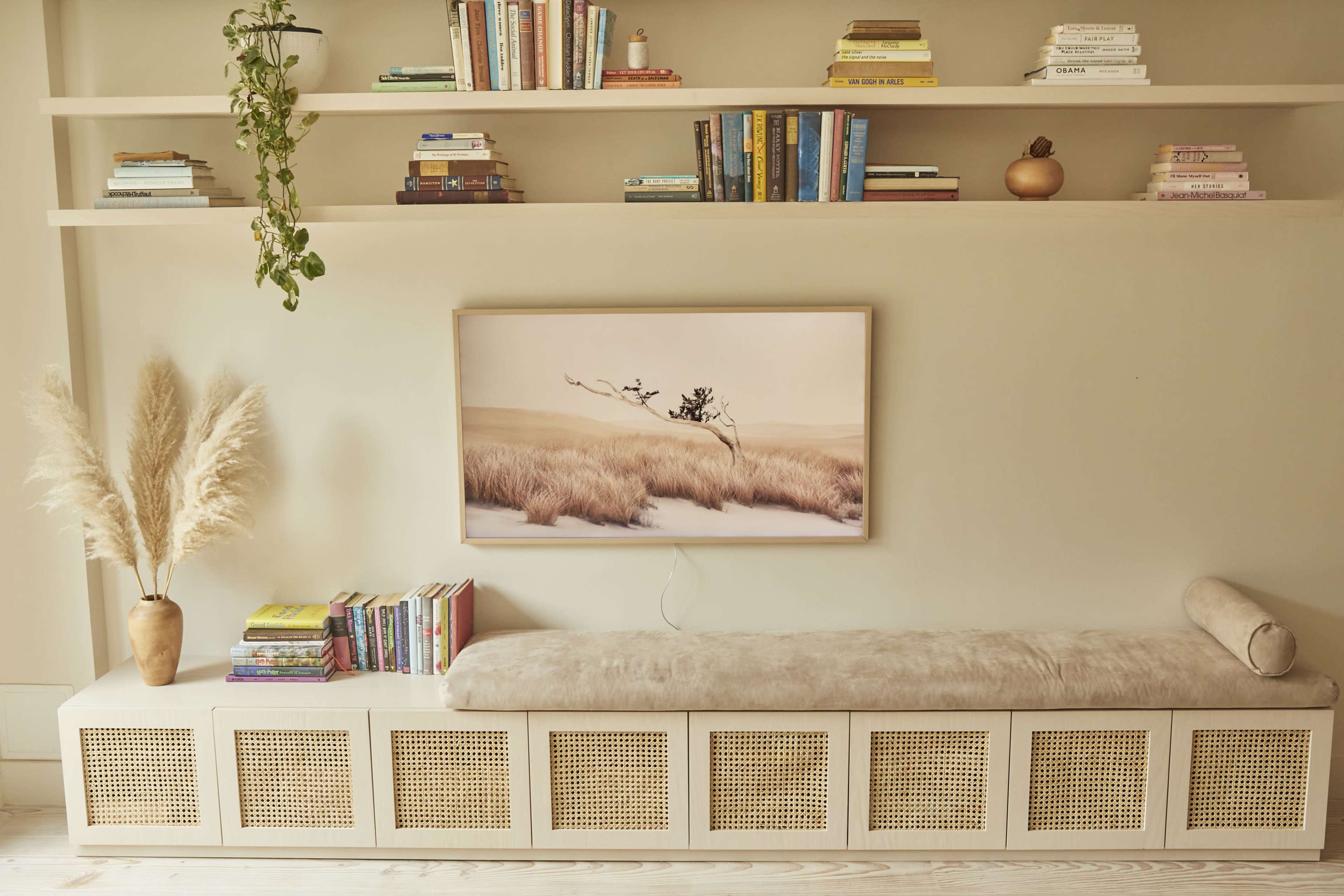 A minimalist living space featuring a wall-mounted shelf with books, a framed landscape artwork, and a padded bench with decorative items and a plant beside it.