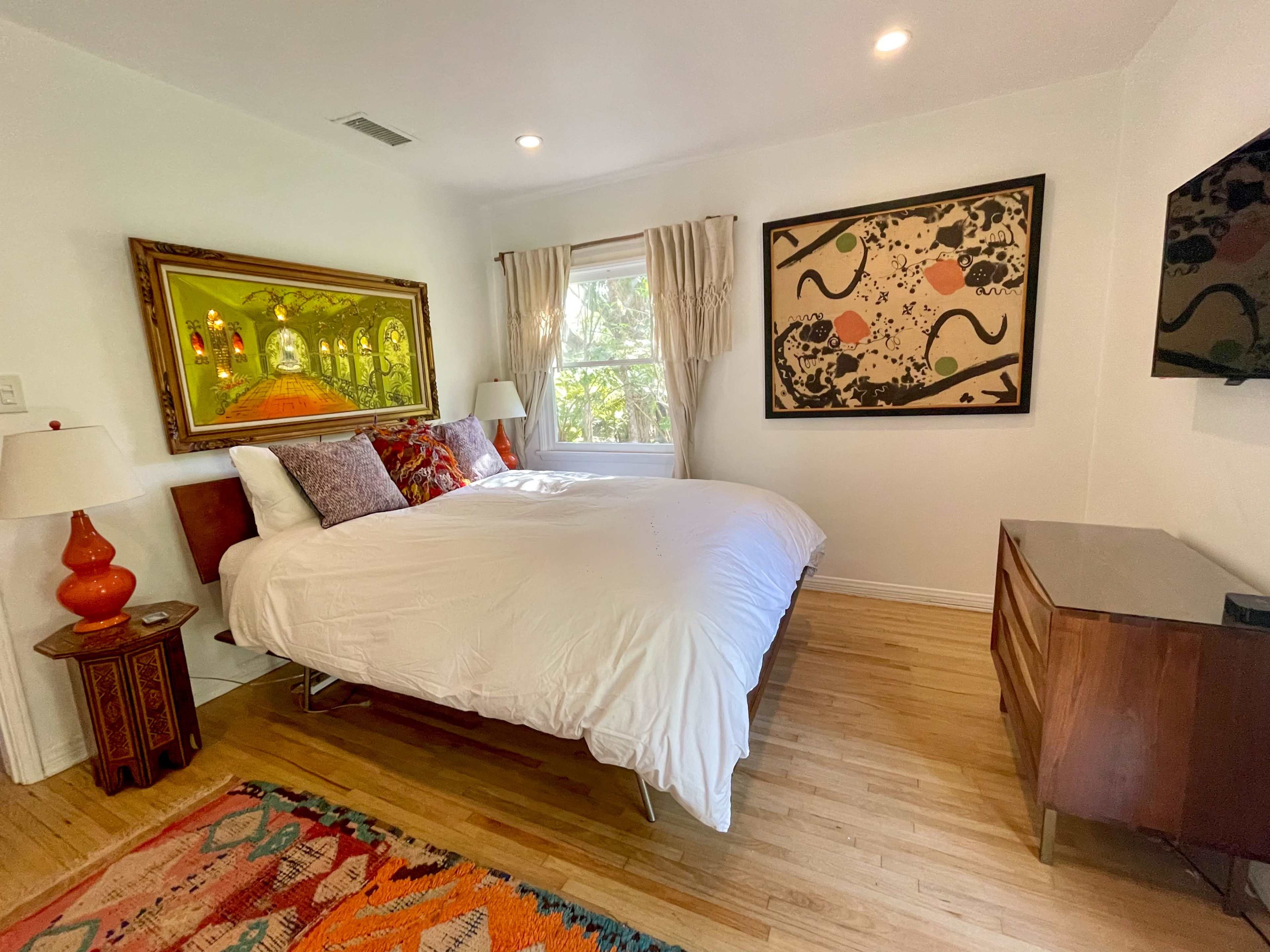 A bright bedroom with a large bed, two side lamps, colorful artwork on the walls, and a wooden dresser.
