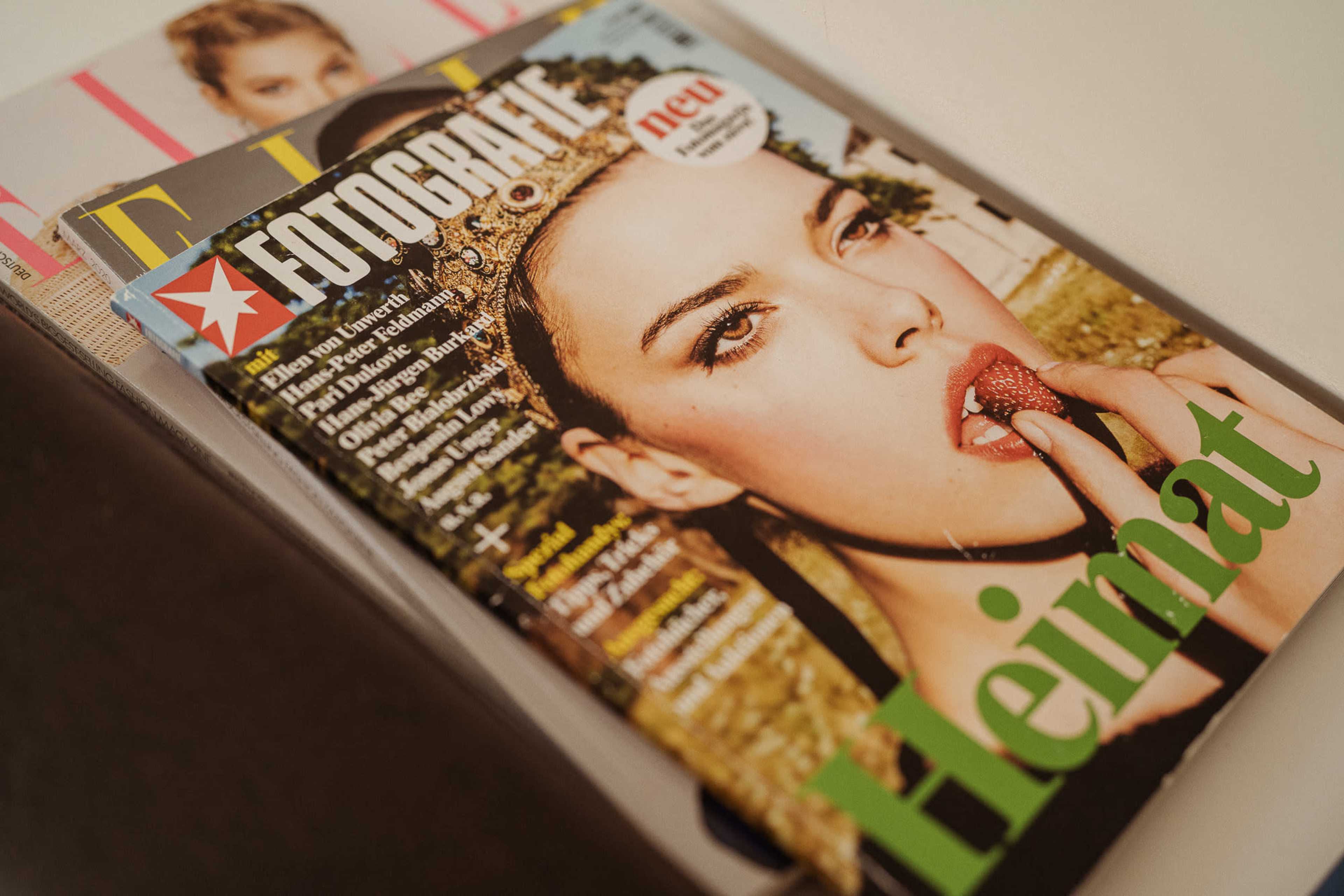 A stack of magazines is shown, with a prominent cover featuring a woman holding a strawberry and the word "Heimat" in green.