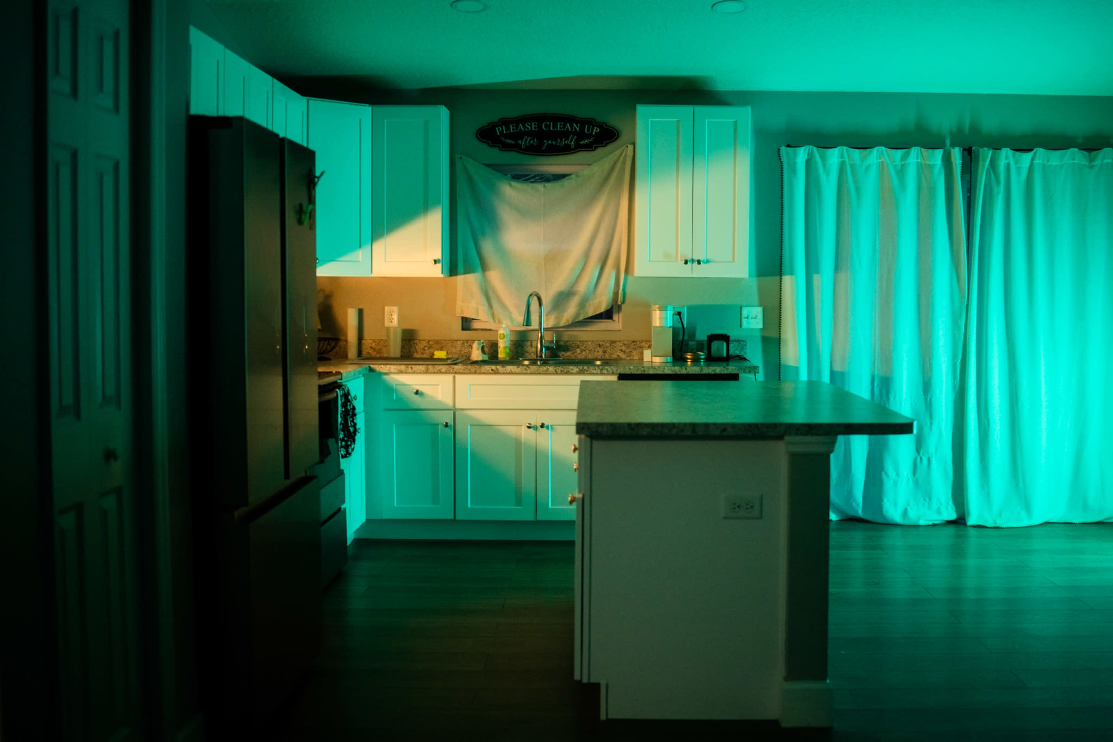 A kitchen with white cabinets and a countertop, illuminated by green light, features a towel hanging in the window and curtains partially drawn.
