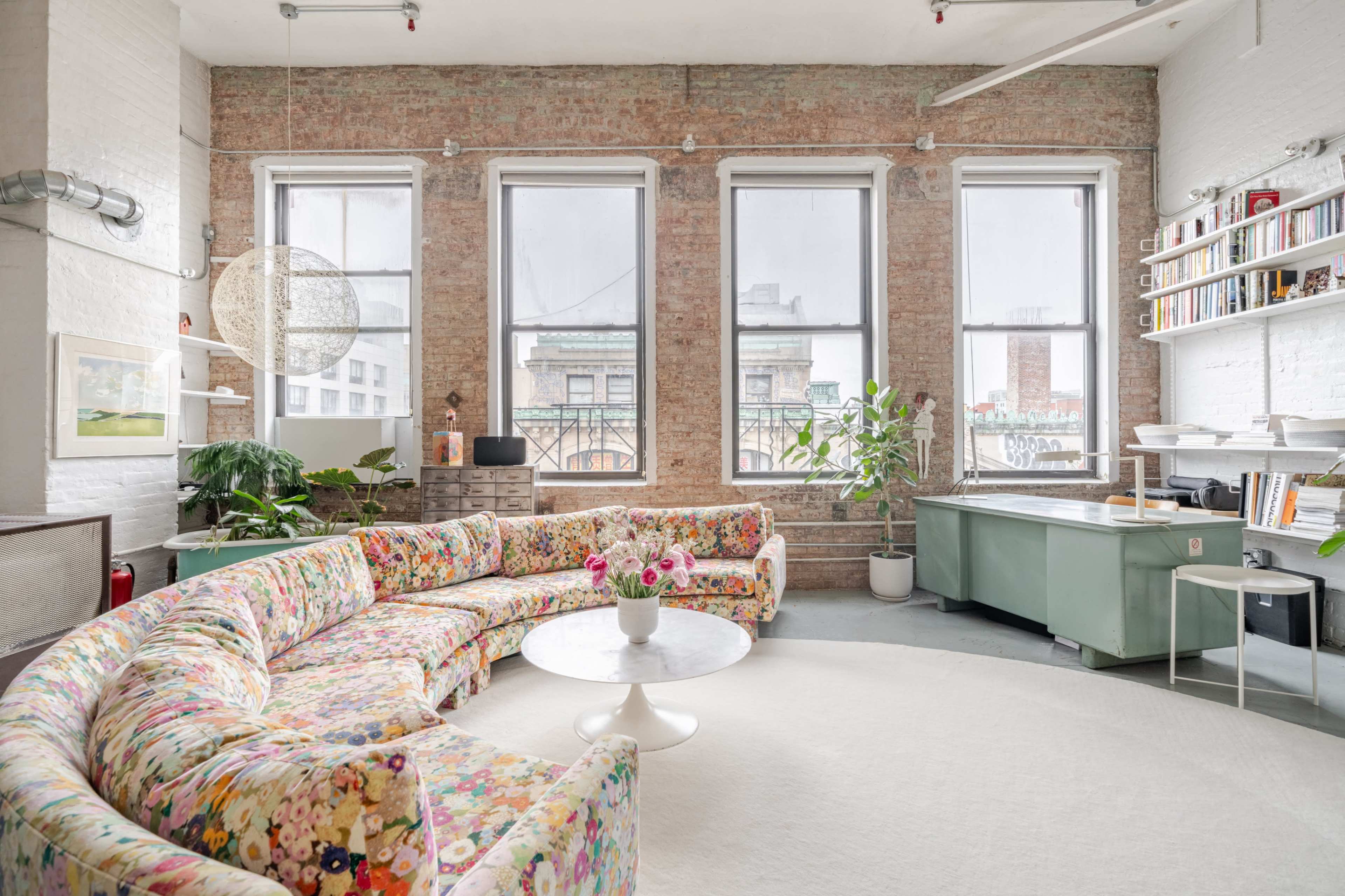 The image shows a spacious, brightly lit room featuring a large floral-patterned sectional sofa, a round coffee table, and a desk against a backdrop of exposed brick walls and large windows.