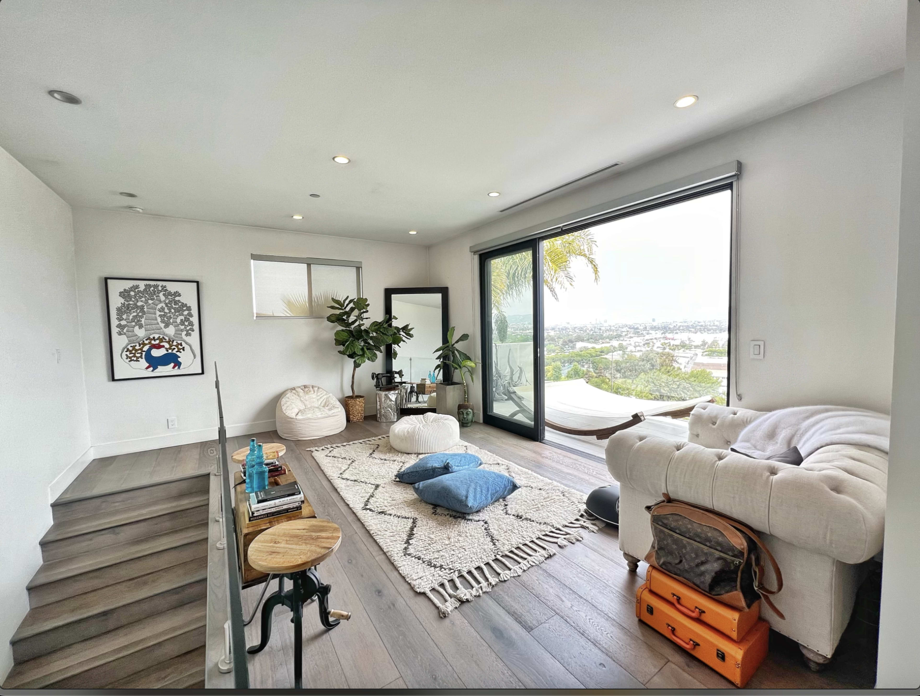 A modern living room features a white couch, oversized cushions, a patterned rug, and large windows overlooking a view.