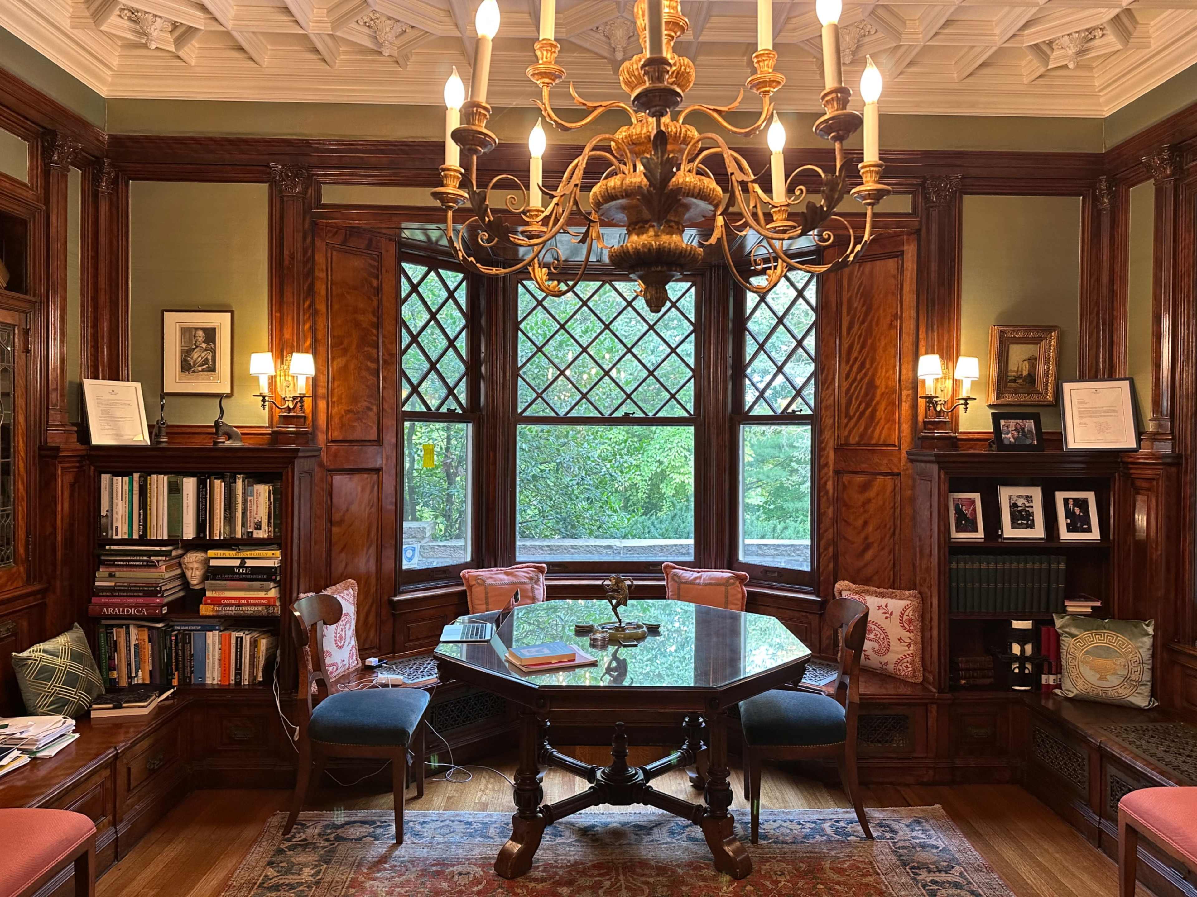 The image shows a well-furnished library with a large wooden table, several chairs, bookshelves filled with books, and a prominent chandelier.