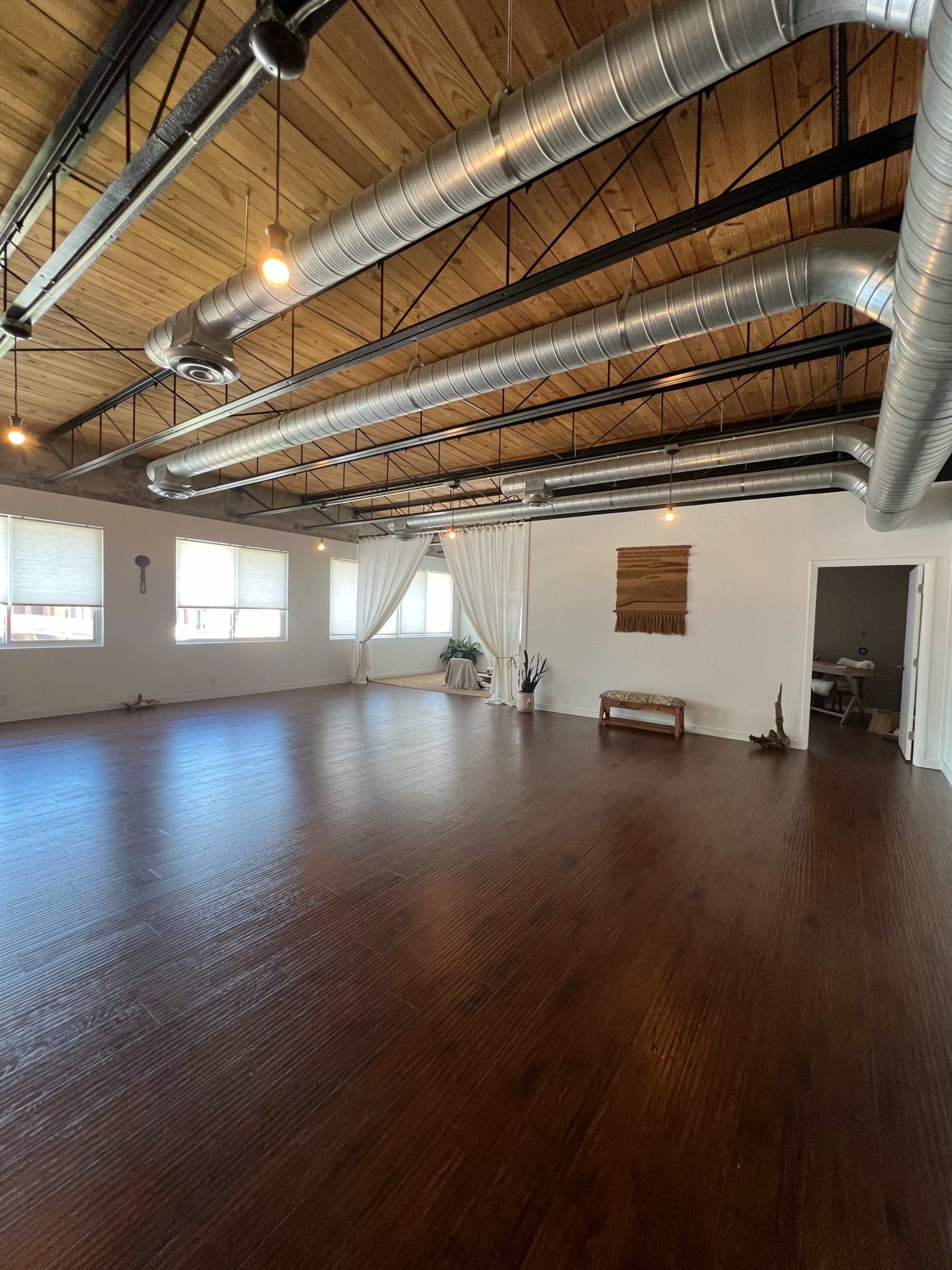 A spacious, well-lit room with wooden flooring, exposed metal ductwork, and large windows featuring white curtains.