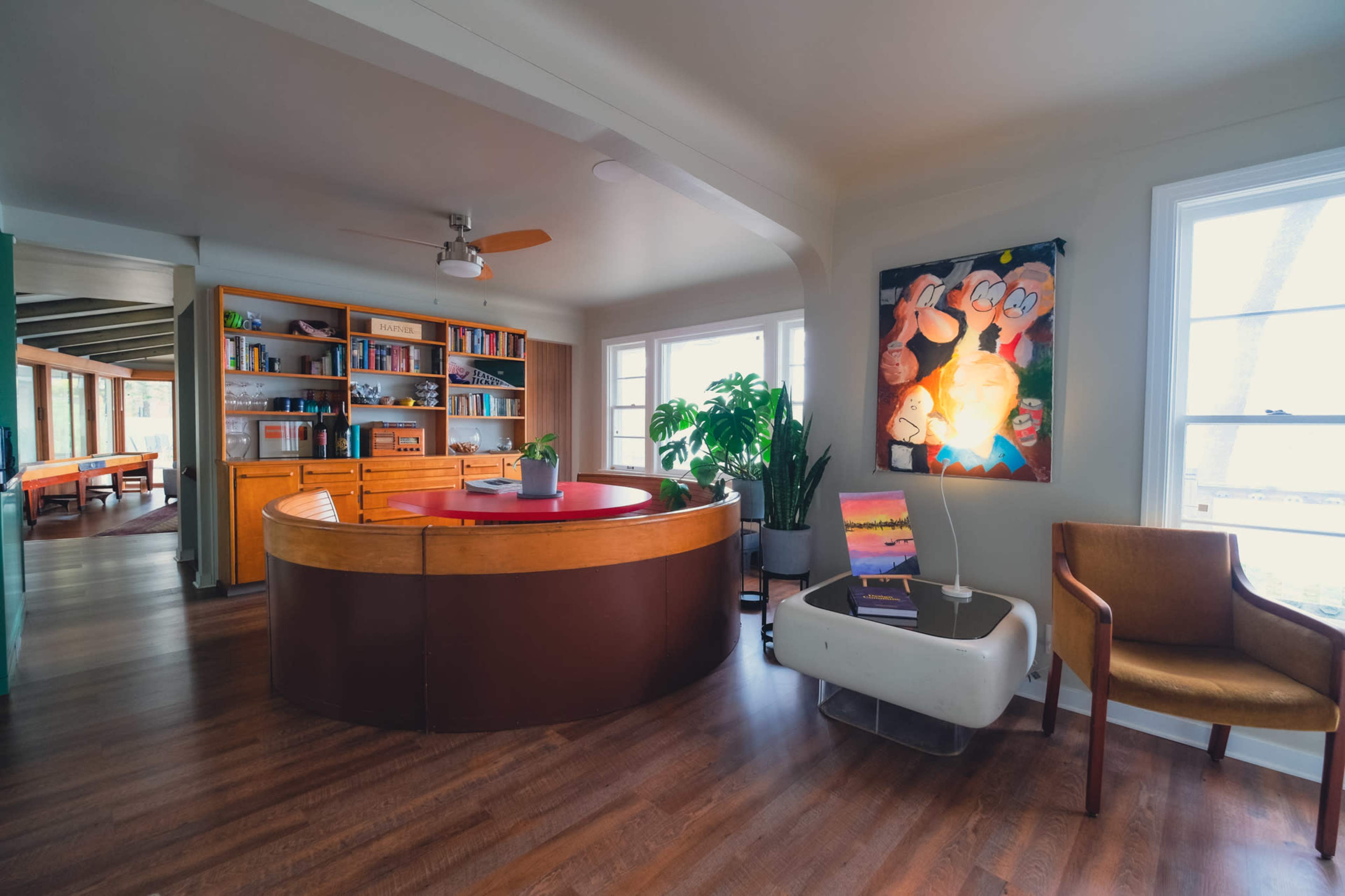 A stylish room featuring a curved wooden reception desk, bookshelves, a large plant, a seating area, and a colorful framed artwork on the wall.