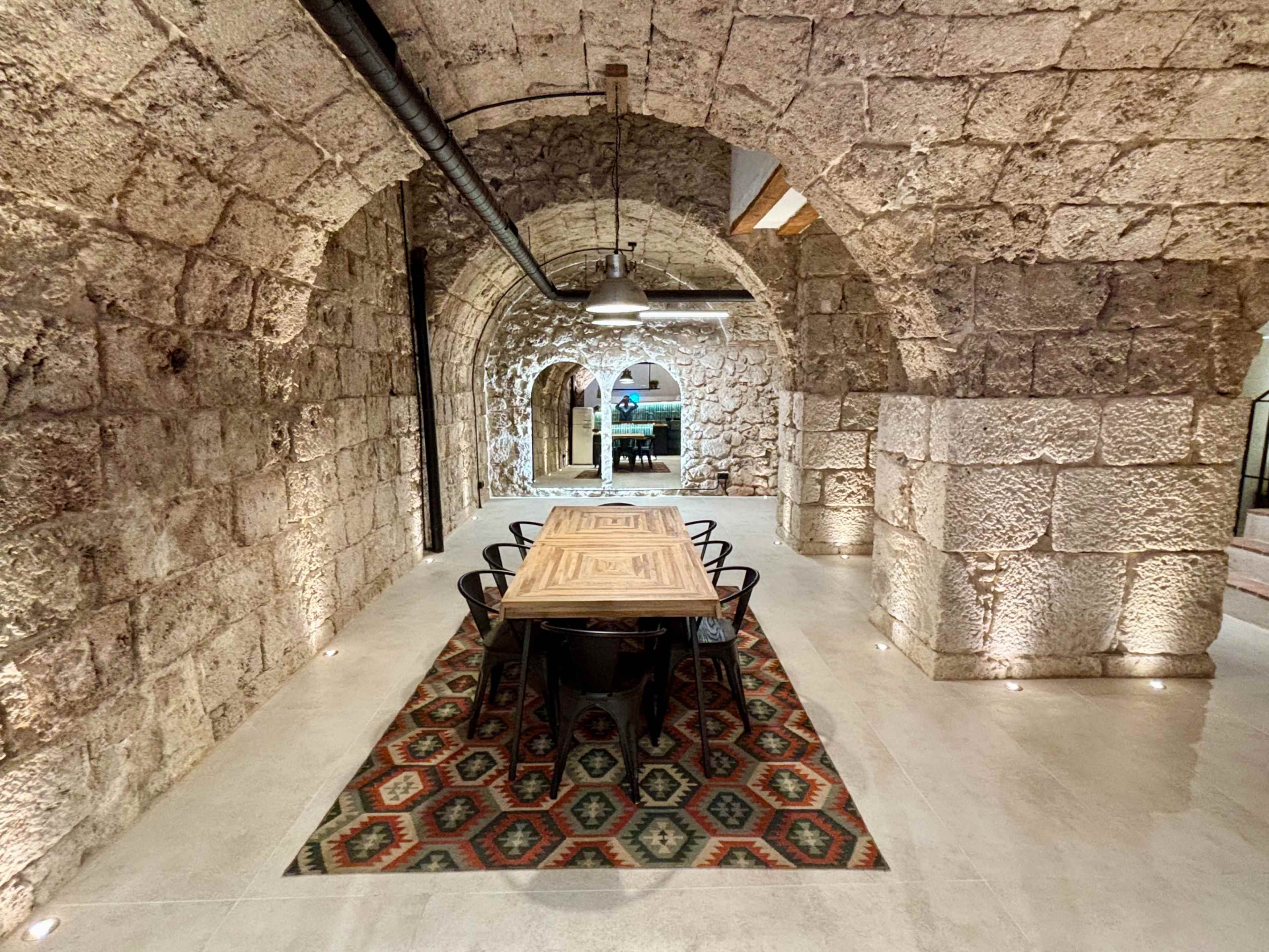 The image shows a long, arched stone hallway with a large wooden table and black chairs in the center, illuminated by soft lighting.