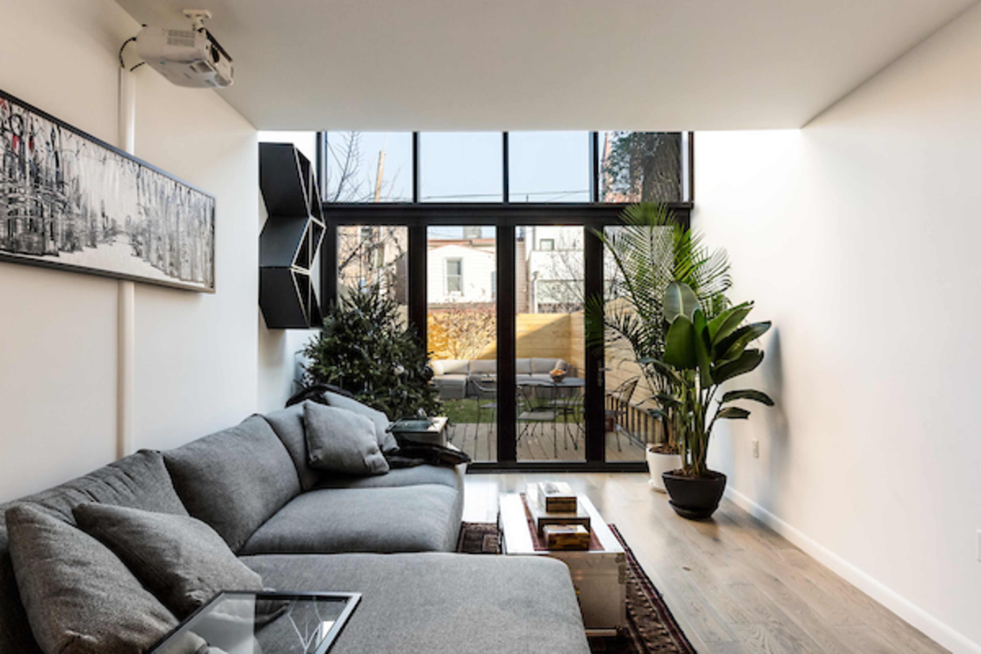 A modern living room featuring a gray sectional sofa, a glass coffee table, and large windows that open to a small outdoor space with greenery.