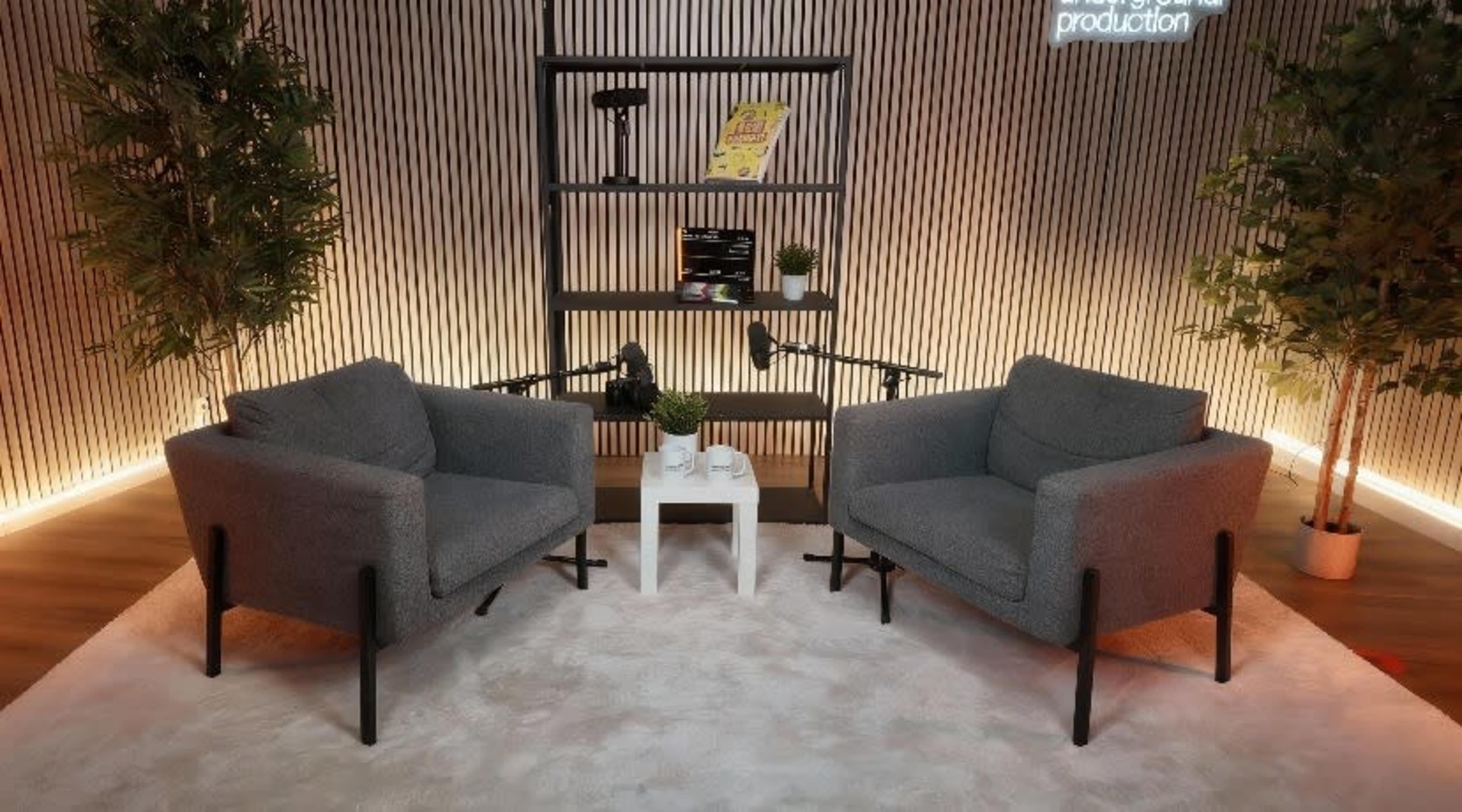 The image shows a modern seating area with two gray armchairs, a small white table, and decorative plants against a textured wall with soft lighting.