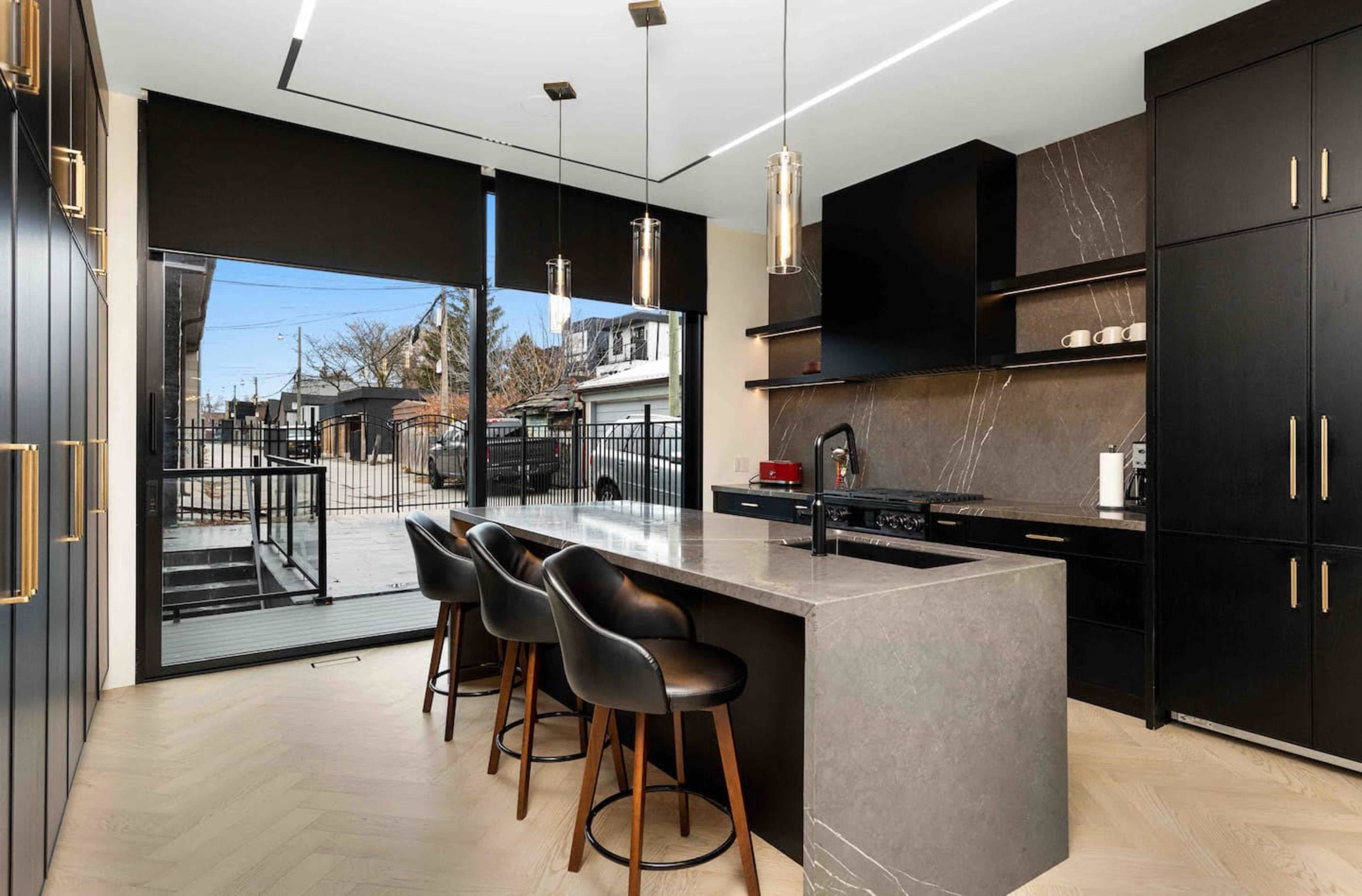 The image depicts a modern kitchen with dark cabinetry, a large island featuring bar stools, and large windows overlooking an outdoor space.