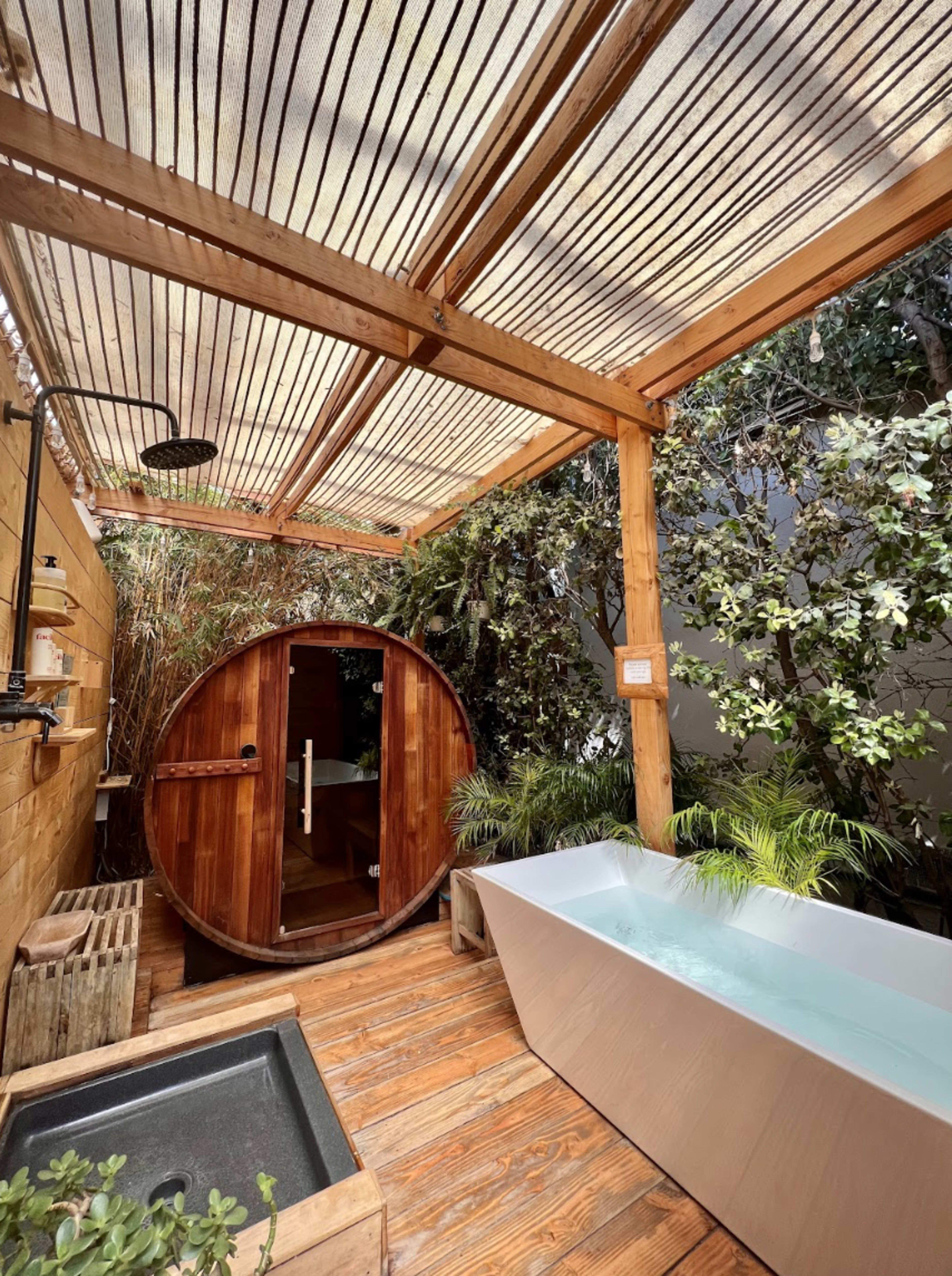 A wooden outdoor spa area featuring a freestanding bathtub, a round wooden sauna, and greenery surrounding the space under a slatted roof.