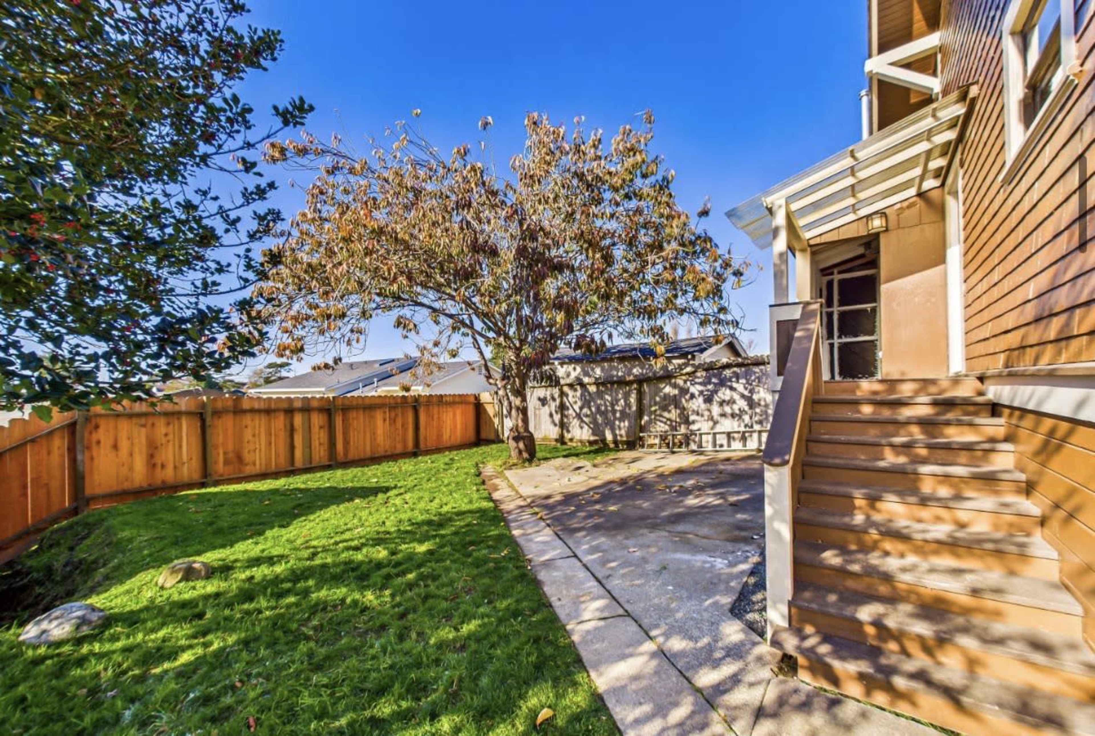 The image shows a side yard with a tree, a grassy area, and a pathway leading to a set of stairs beside a house, enclosed by a wooden fence.