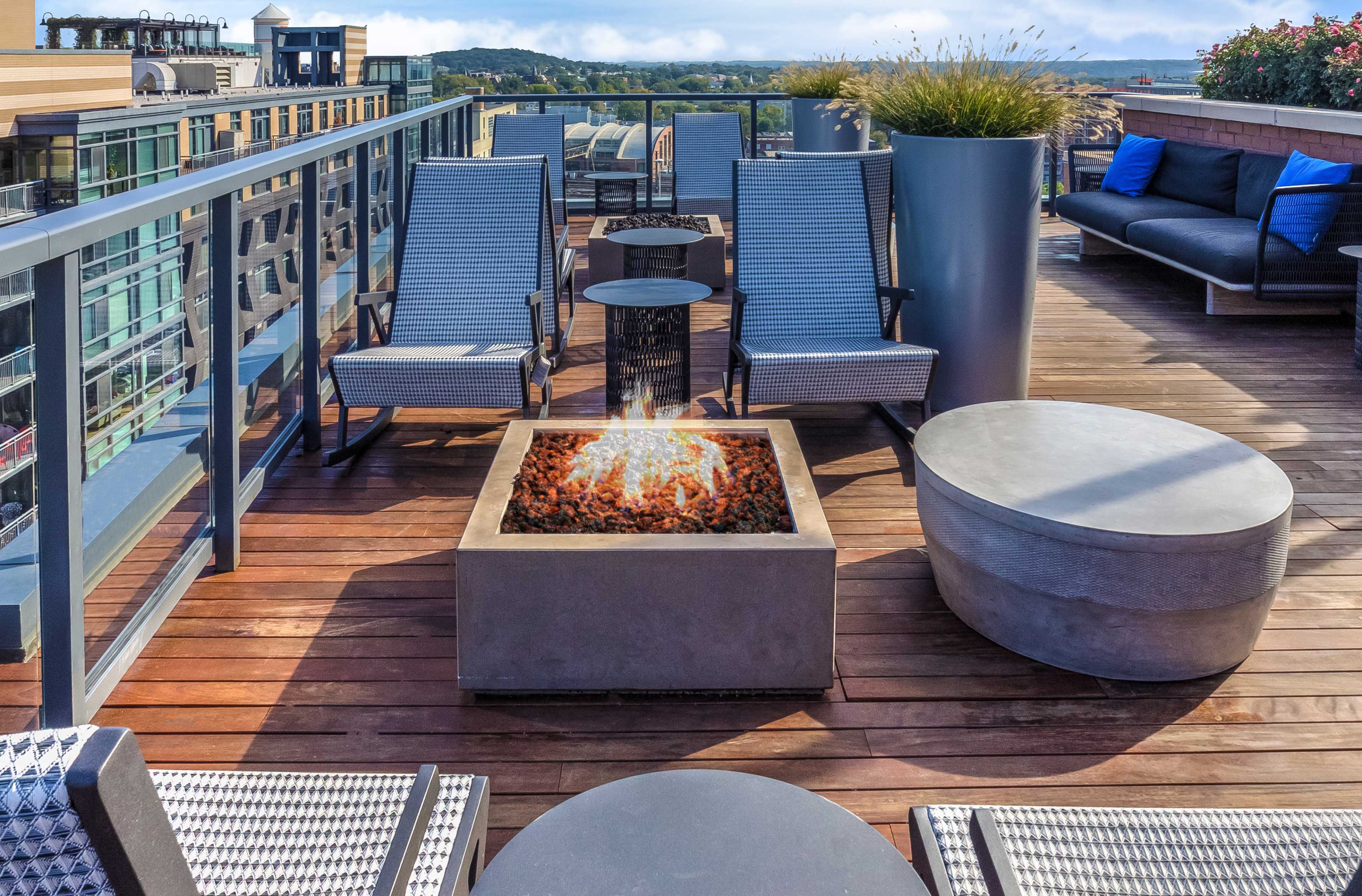A rooftop terrace with lounge chairs arranged around a fire pit, surrounded by planters and offering a view of the landscape beyond.