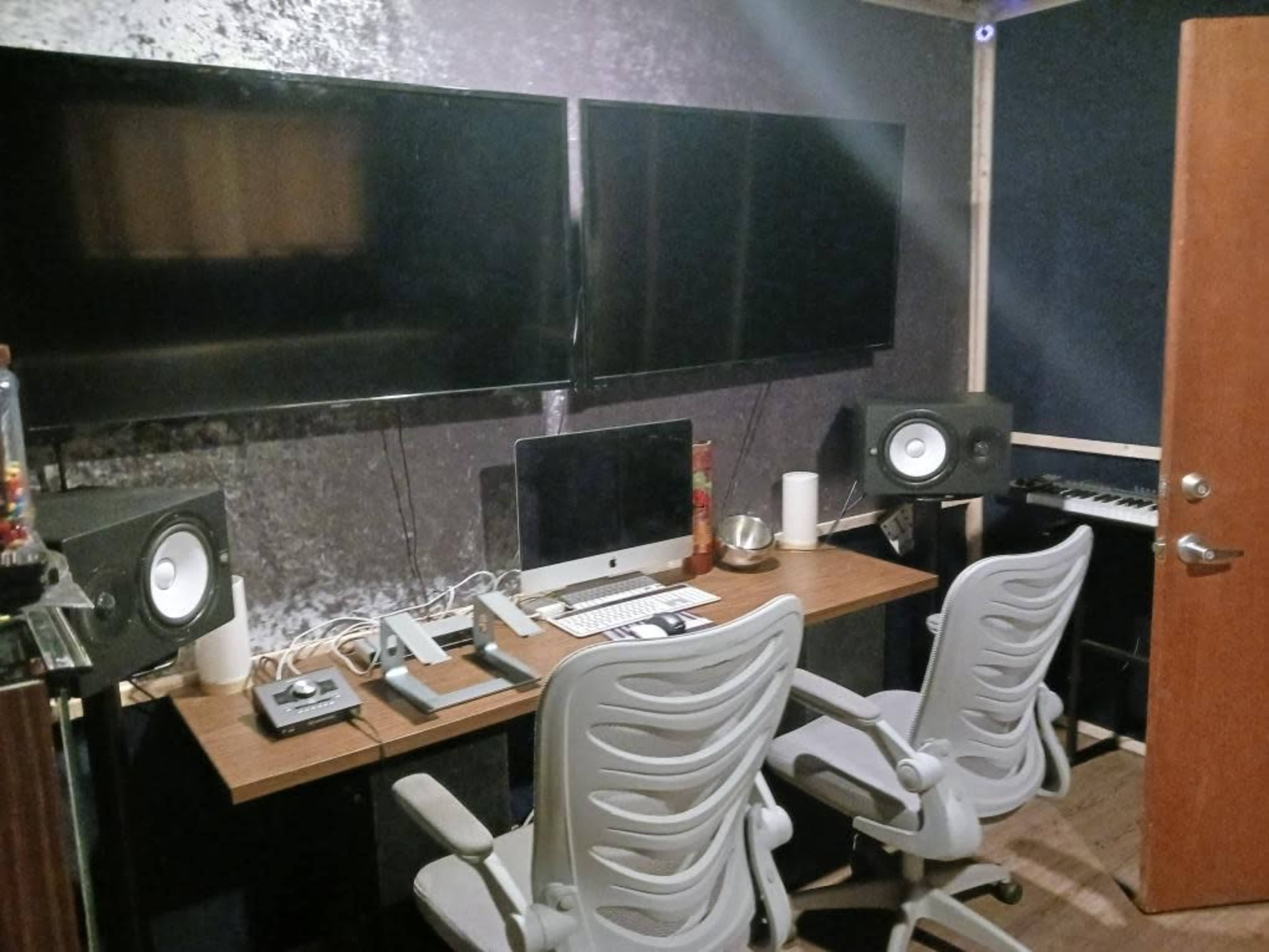 The image shows a modern music studio setup featuring two large monitors, a desktop computer, audio speakers, and a keyboard along with two ergonomic office chairs.