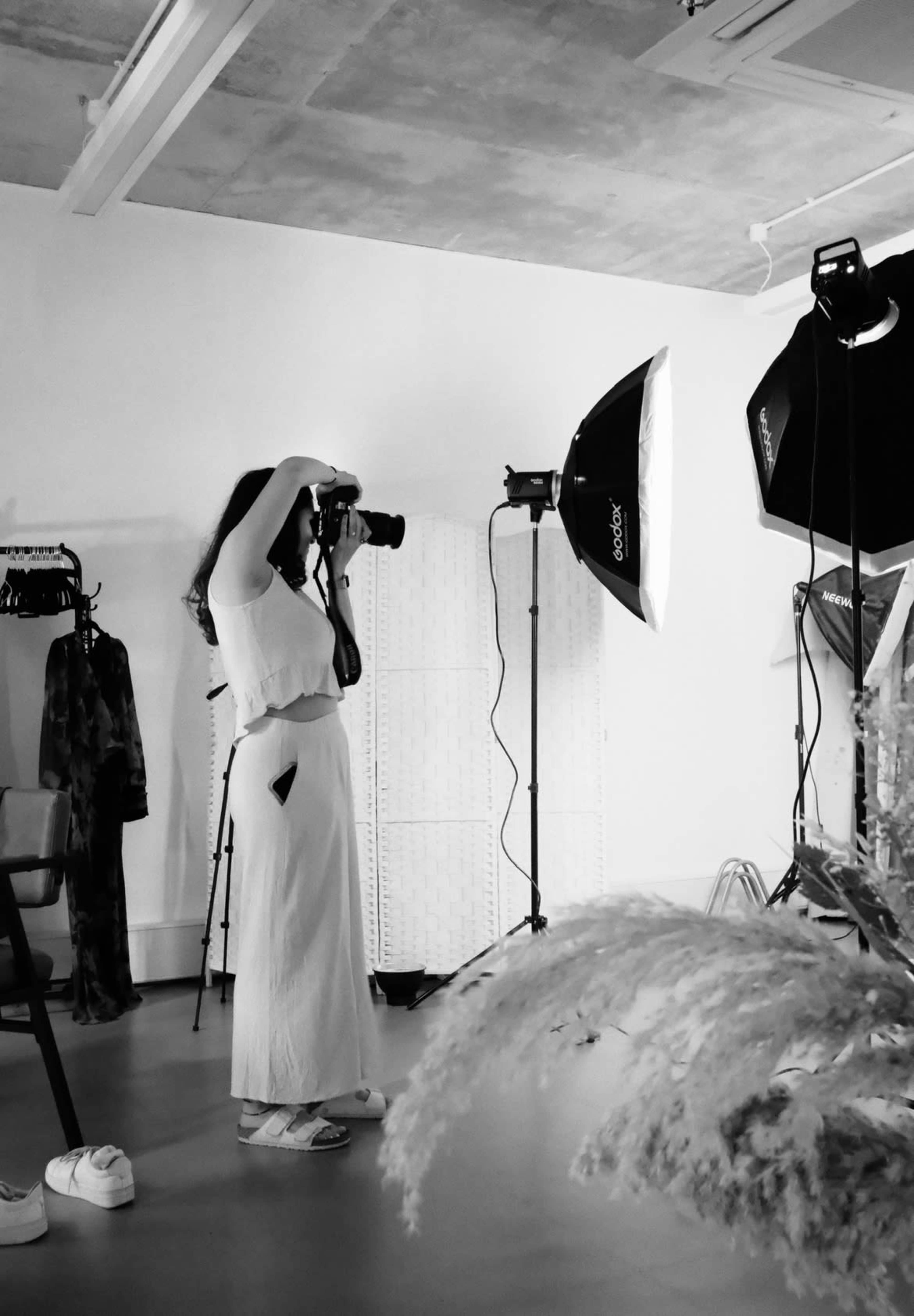 A woman in a white outfit is standing in a studio, holding a camera and photographing a setup with studio lights.