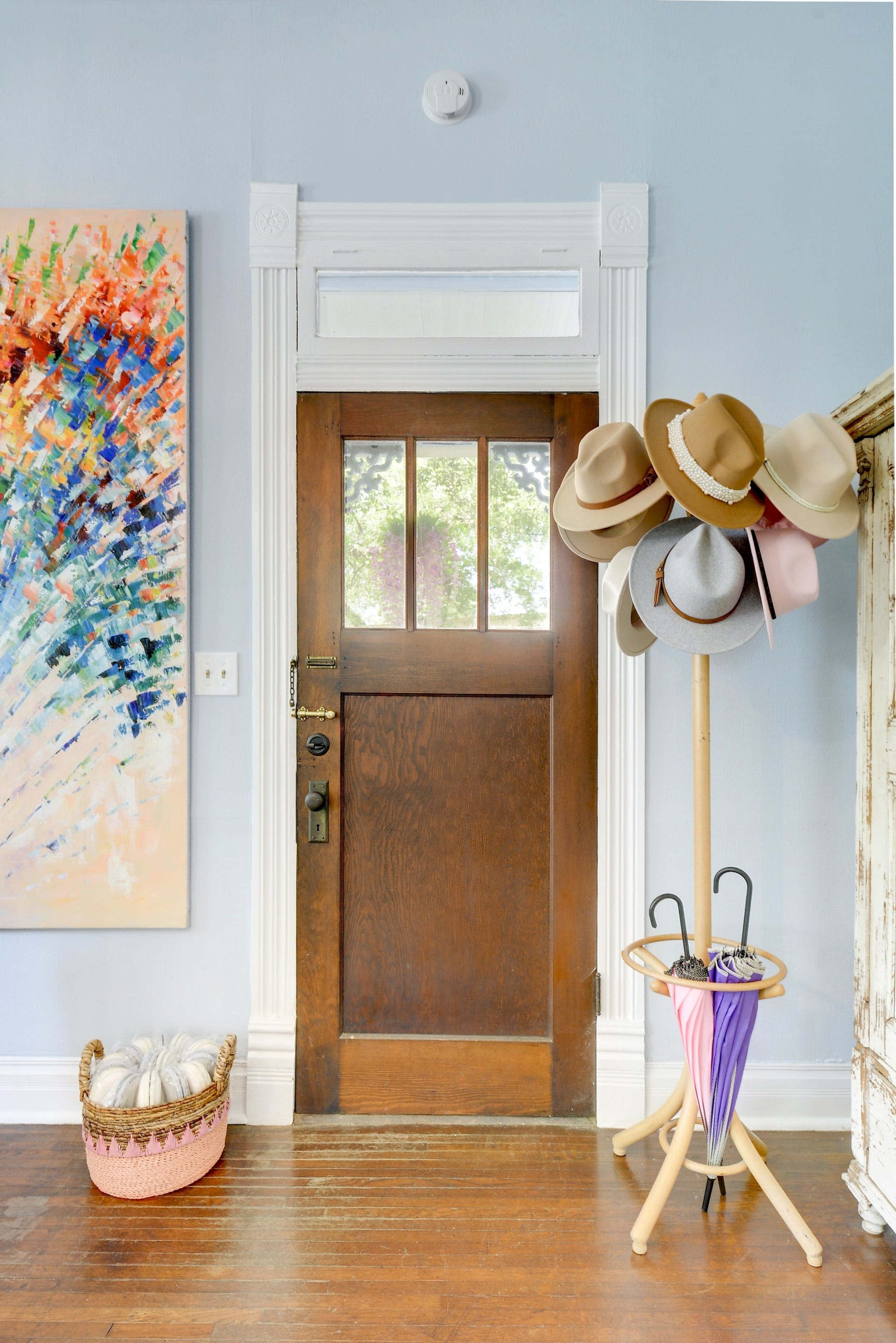 An entrance area featuring a brown wooden door, a colorful abstract painting on the wall, and a hat rack displaying several hats.