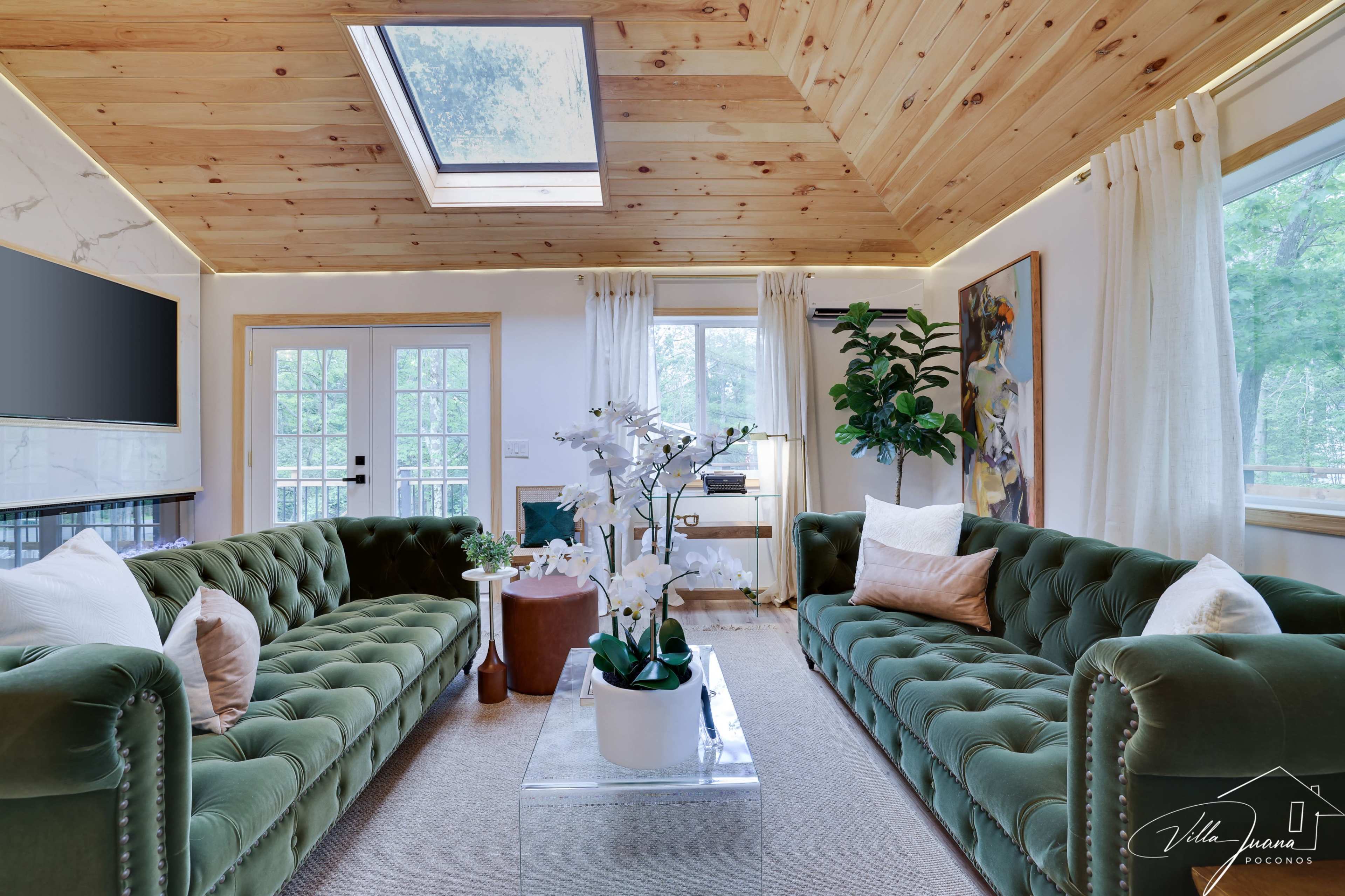 The living room features two plush green velvet sofas facing each other, with a glass coffee table and a large window allowing natural light to enter.