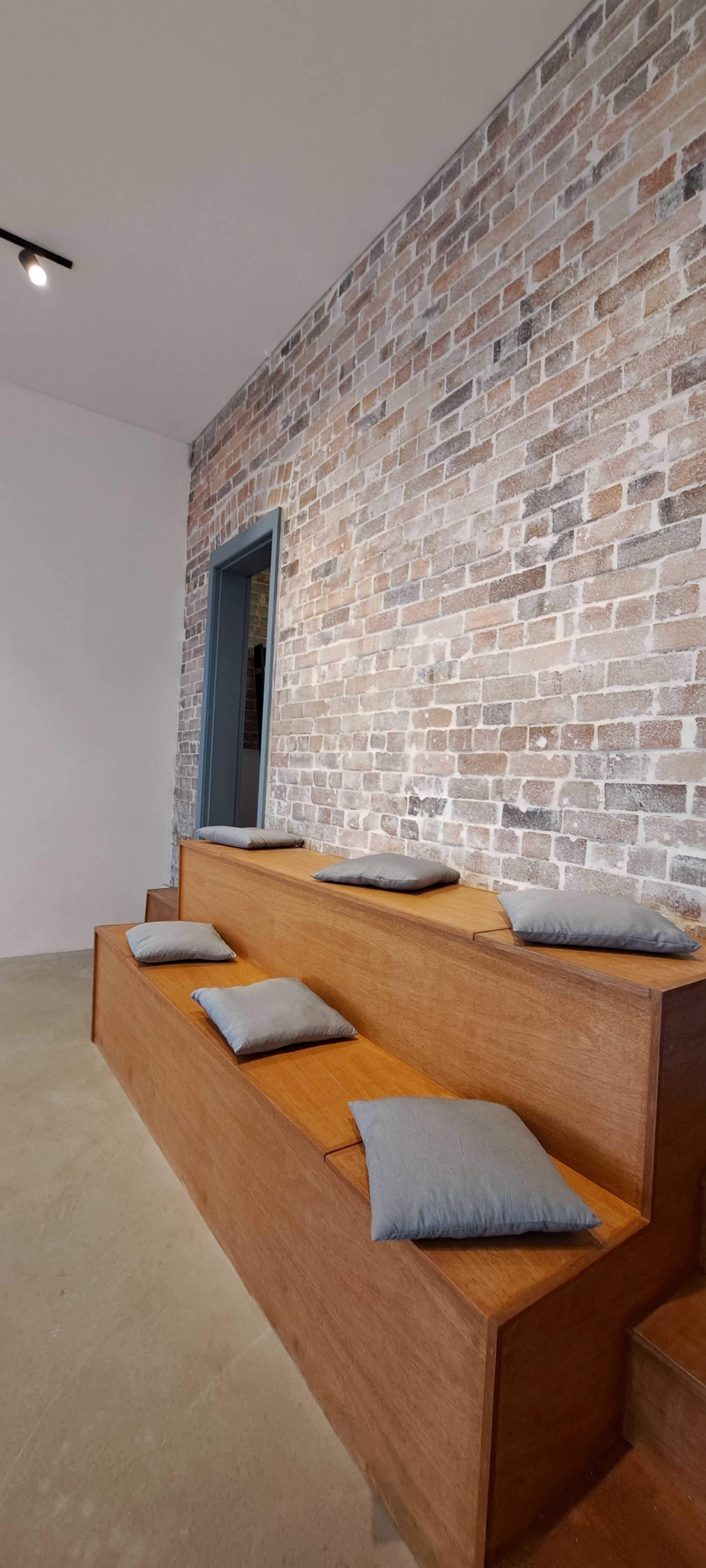 The image shows a minimalist seating area with wooden steps and gray cushions against a textured brick wall.