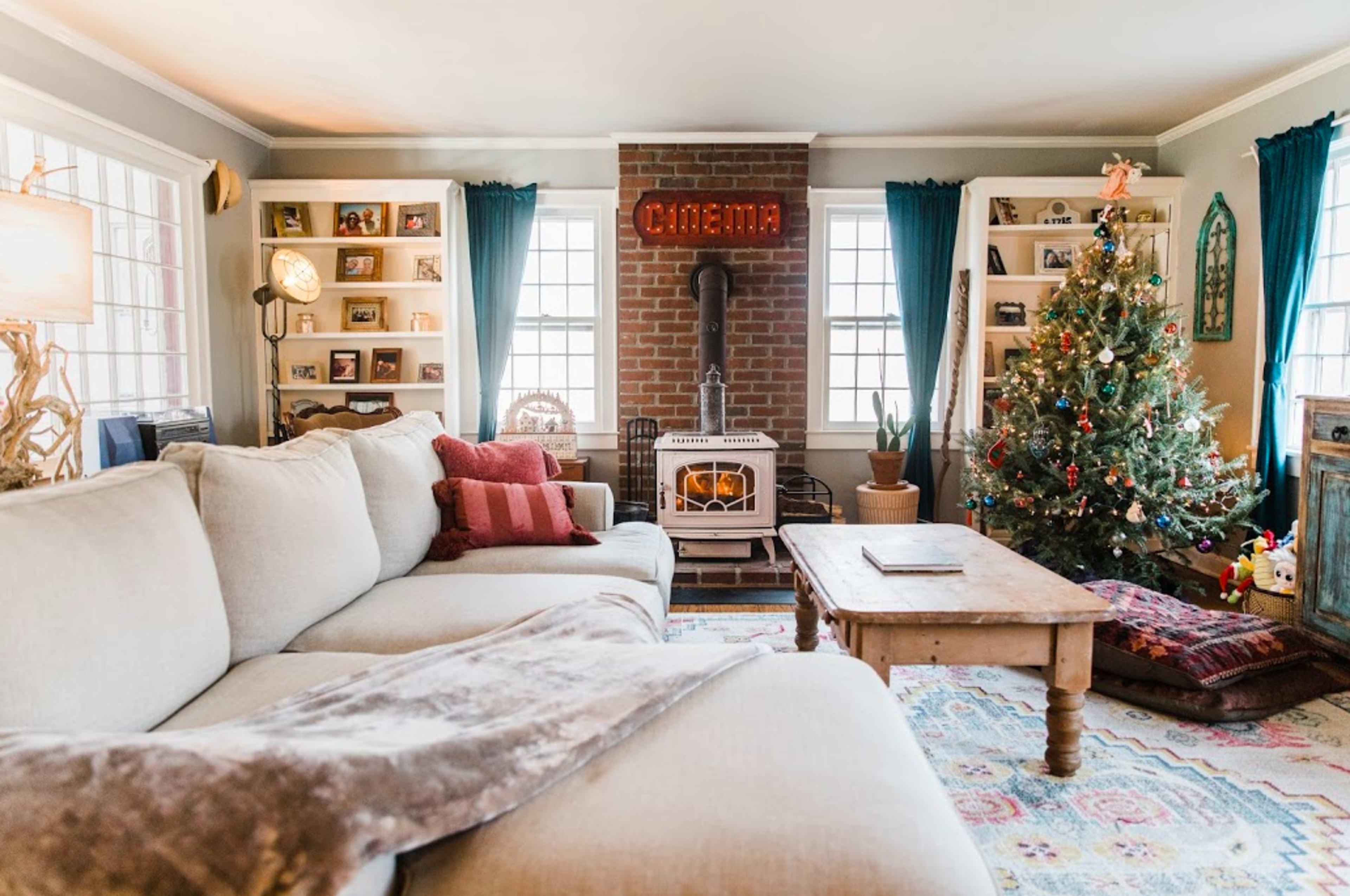 A cozy living room features a large sectional sofa, a brick fireplace with a vintage "Cinema" sign, and a decorated Christmas tree next to a wooden coffee table.