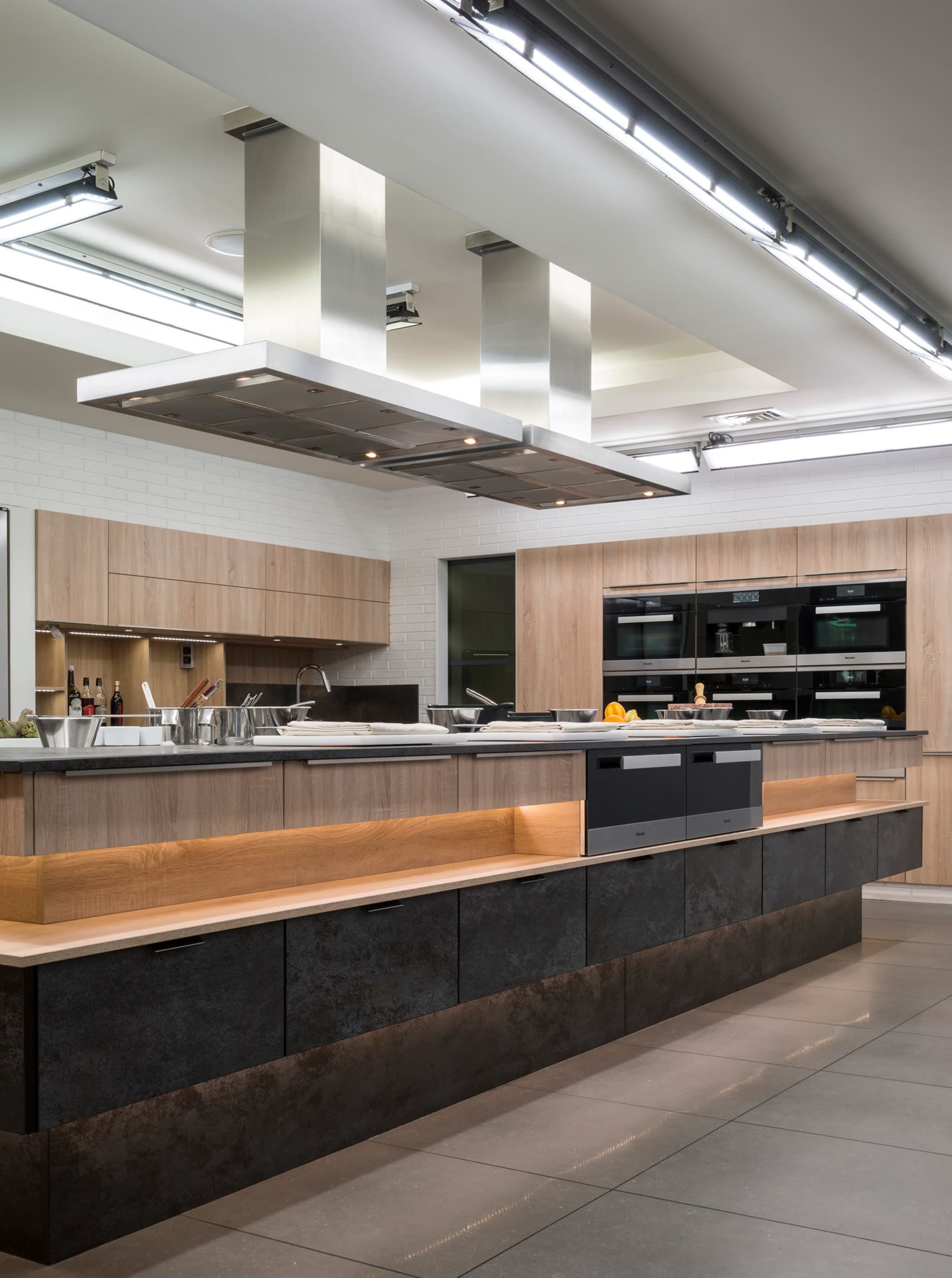 The image shows a modern kitchen with a central island featuring an integrated cooktop, surrounded by wooden cabinetry and built-in appliances.