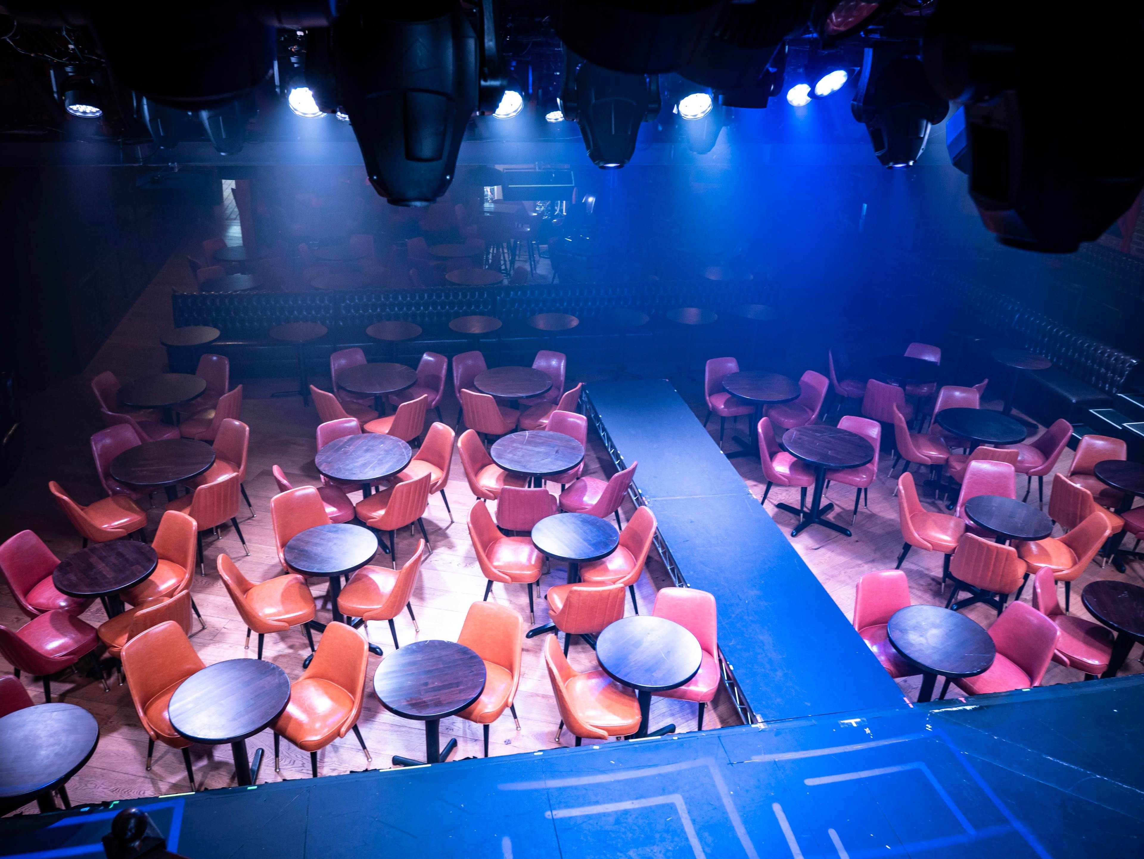 A dimly lit performance venue with rows of round tables and chairs arranged around a central stage.