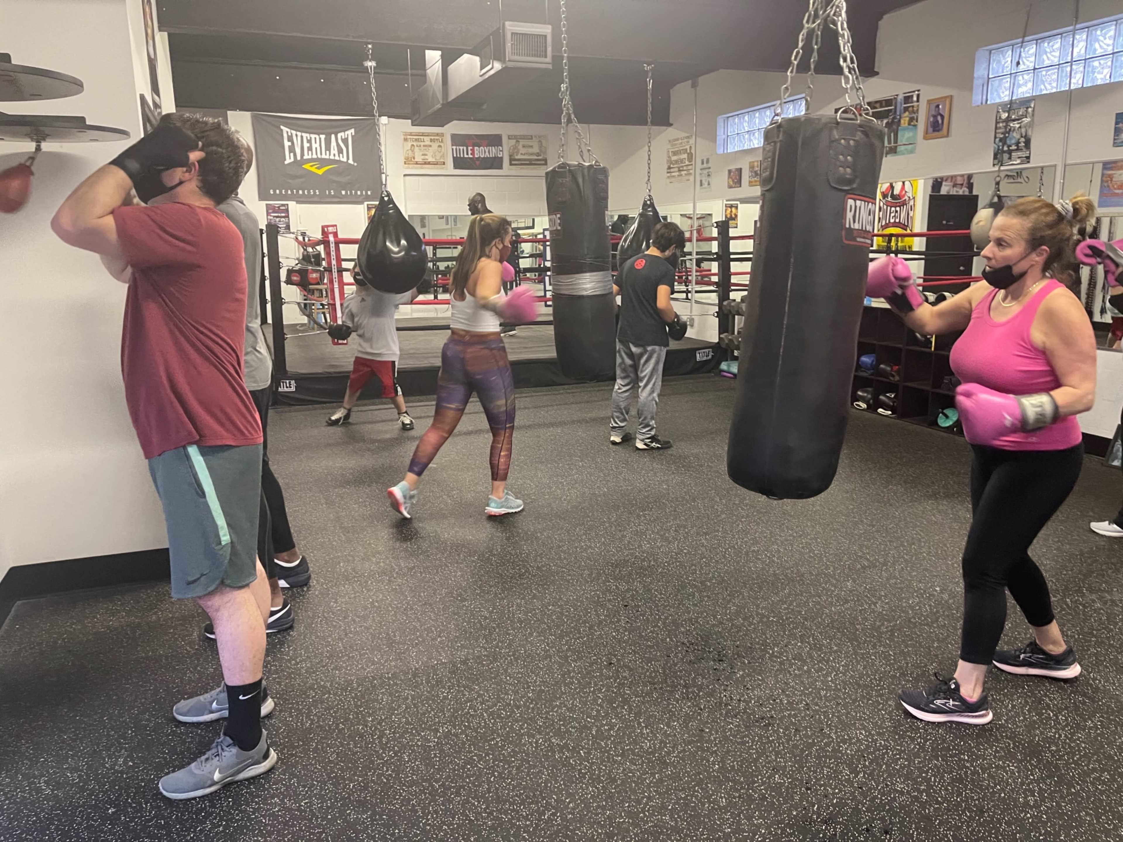 A group of people exercises in a boxing gym, using punching bags and wearing gloves and masks.