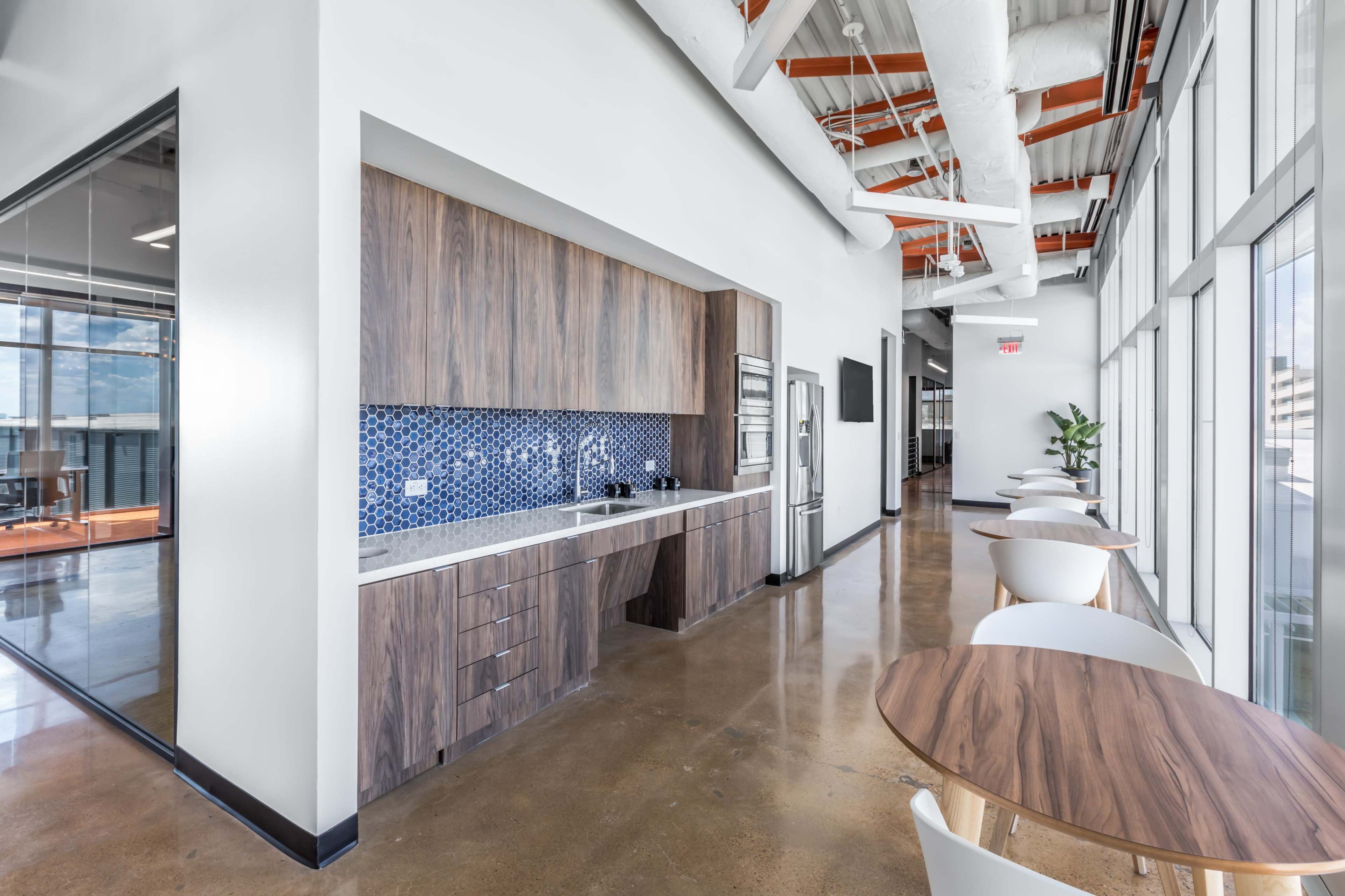 The image shows a modern office kitchen area with wooden cabinetry, blue tiled backsplash, and a long hallway alongside tables and chairs.