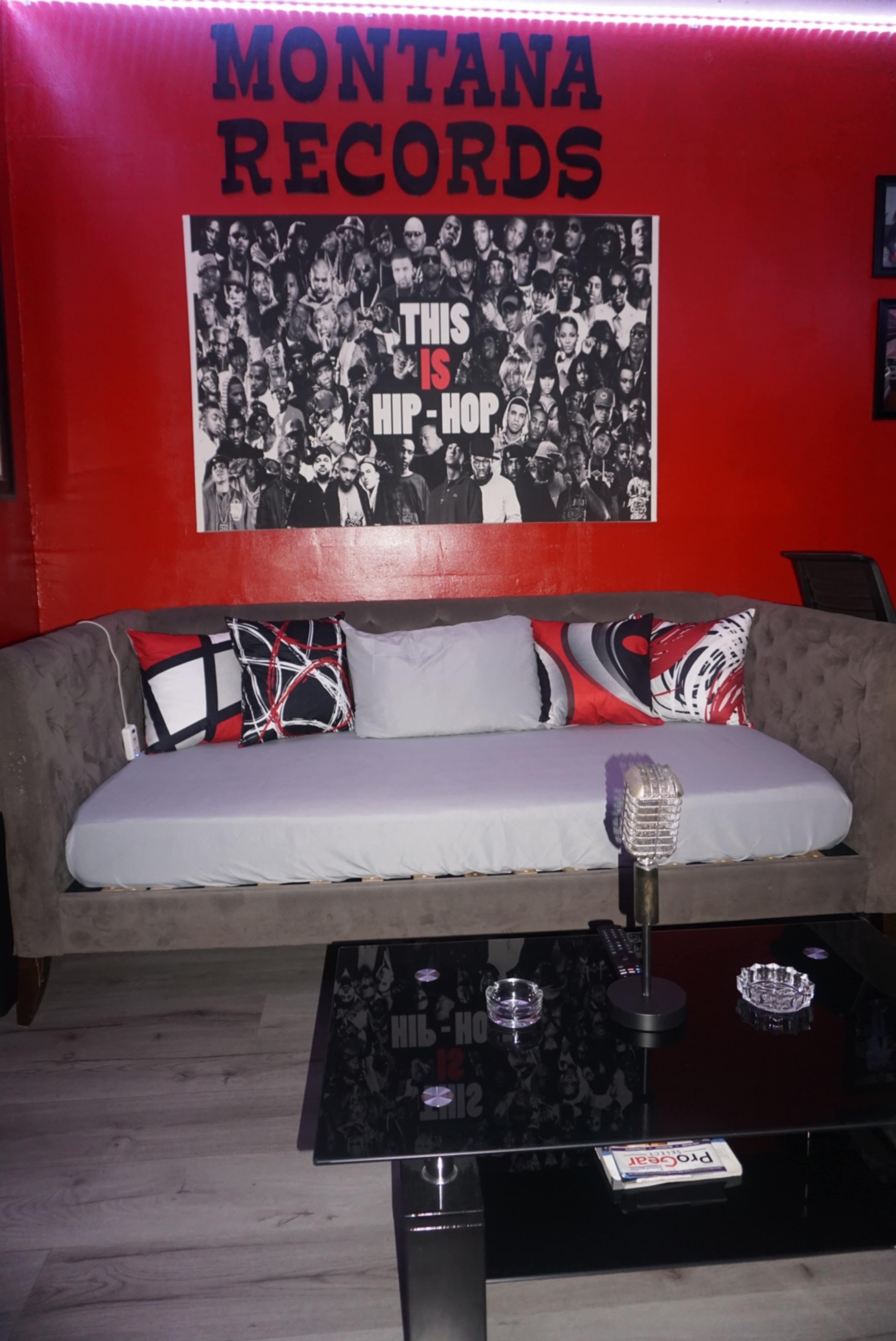 The image shows a modern, red-themed lounge area featuring a plush sofa with decorative pillows, a glass coffee table, and a wall adorned with a large black-and-white graphic of hip-hop artists.