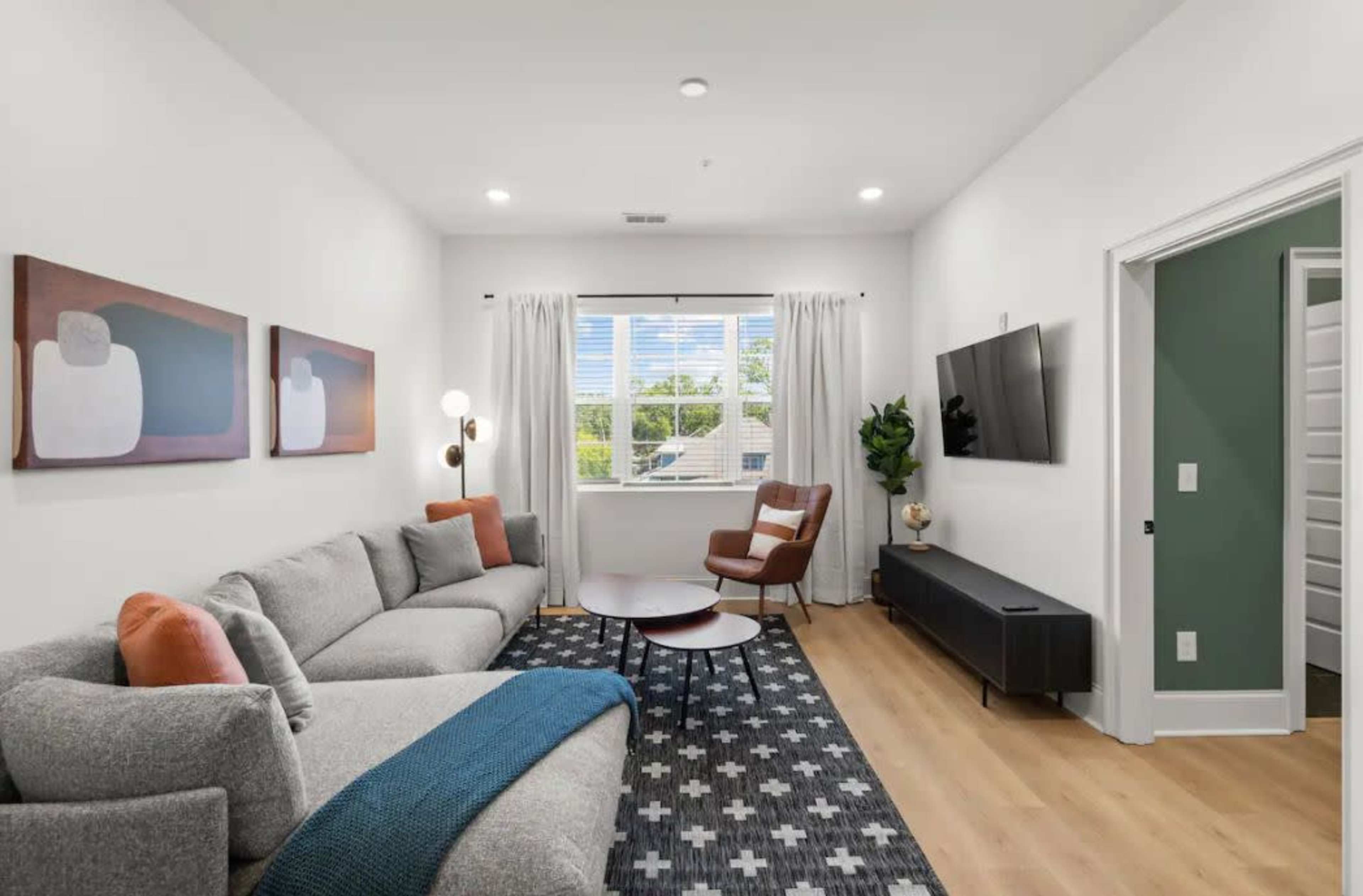 A modern living room features a gray sectional sofa, a round coffee table, and artworks on the walls, with a window offering natural light and a view outside.