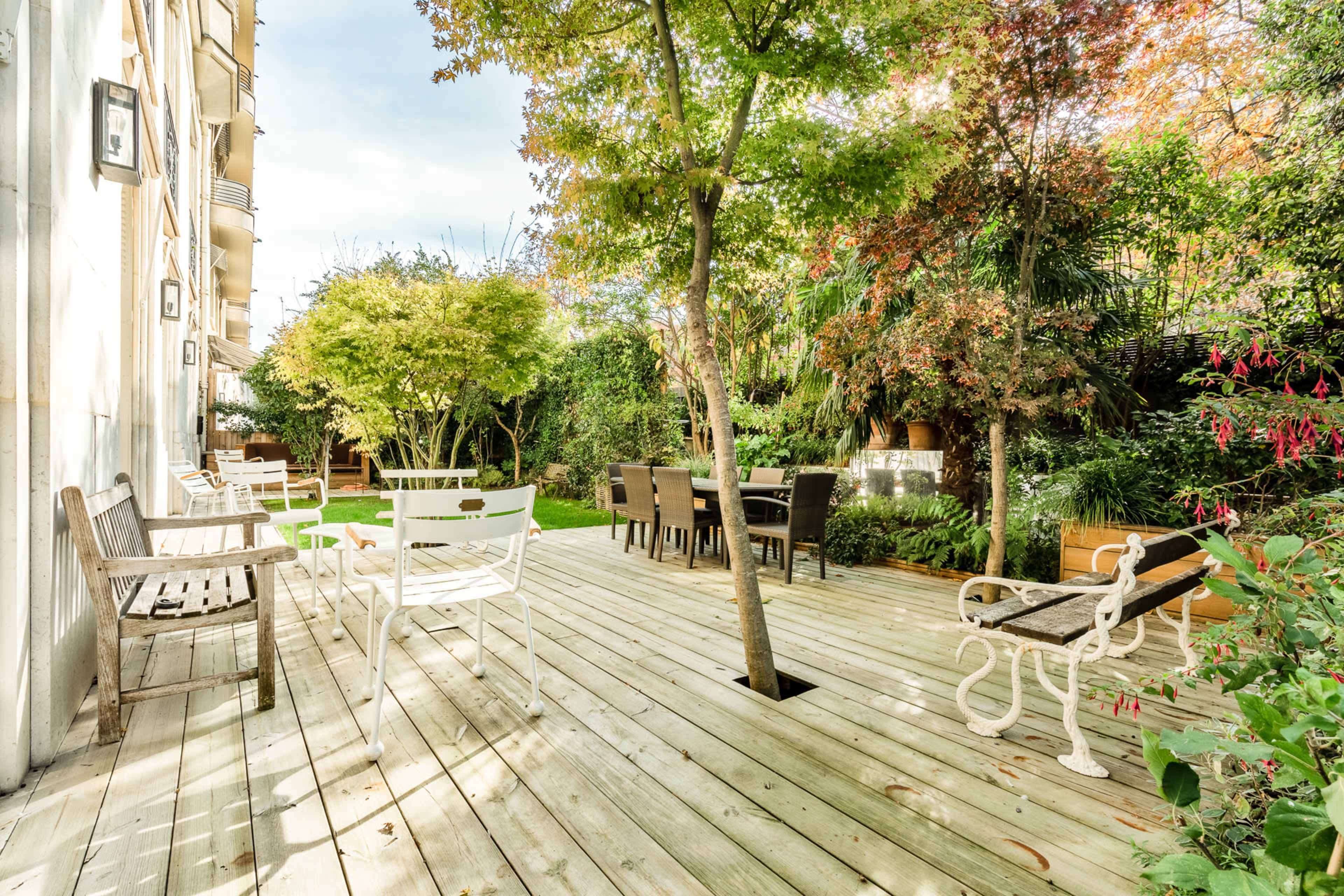 A wooden deck surrounded by greenery, with several seating areas and a dining table under trees.
