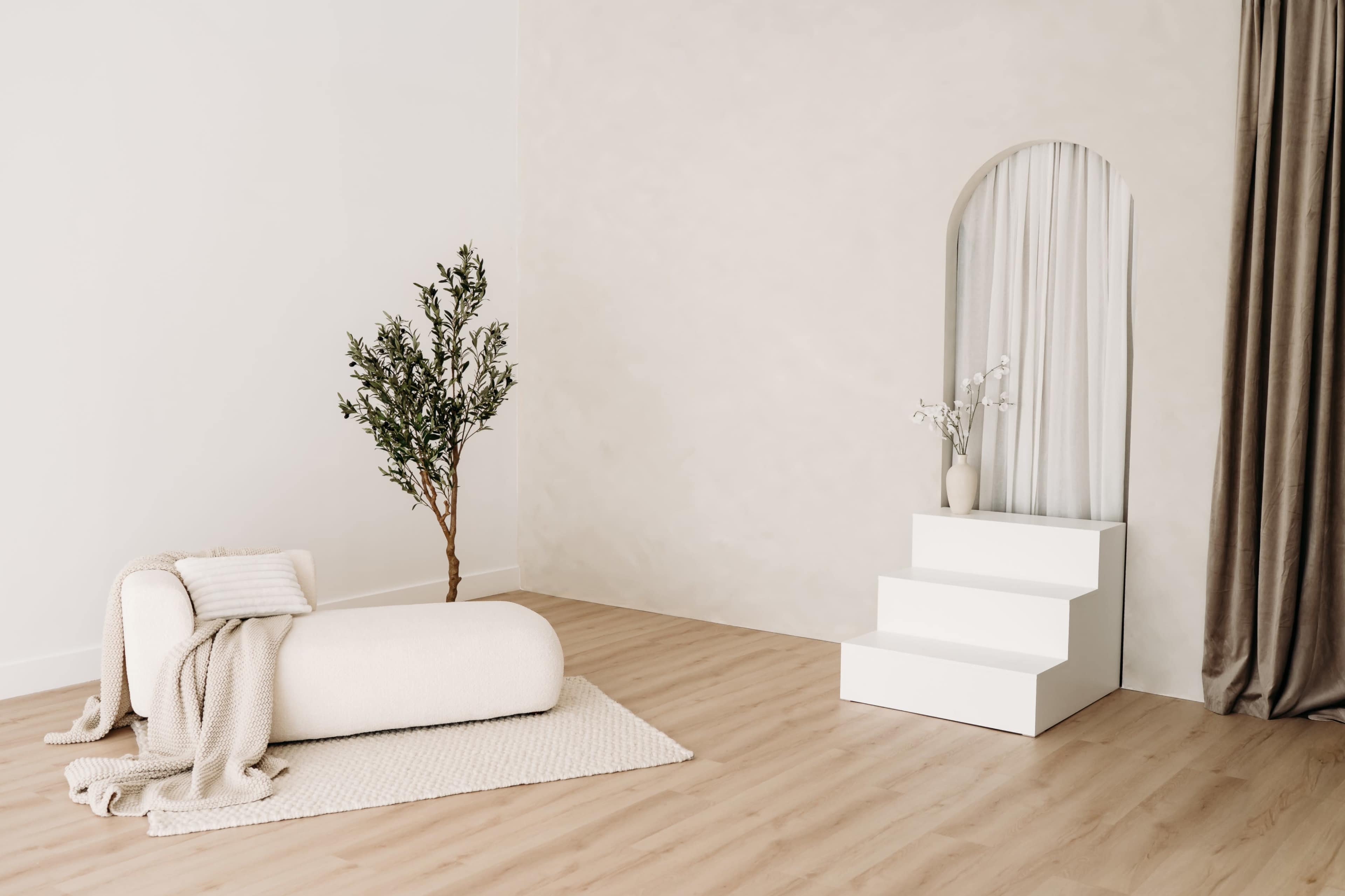 The image features a minimalist interior design with a round couch on a textured rug, a tree, a three-step platform, and a vase with flowers against a light-toned wall.