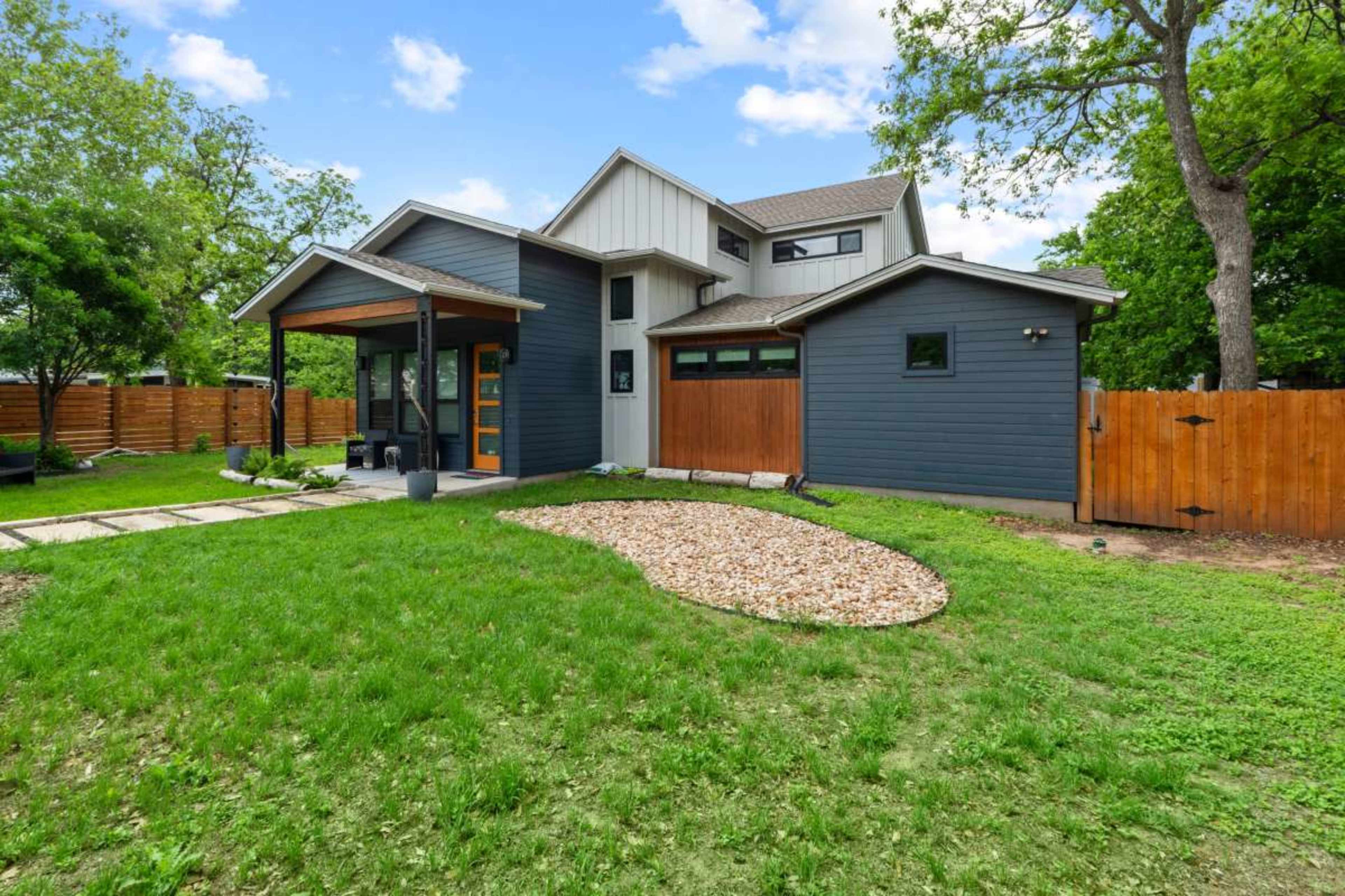 The image shows a modern two-story house with a mix of siding colors, a landscaped yard featuring a gravel circle, and a wooden fence enclosing the property.