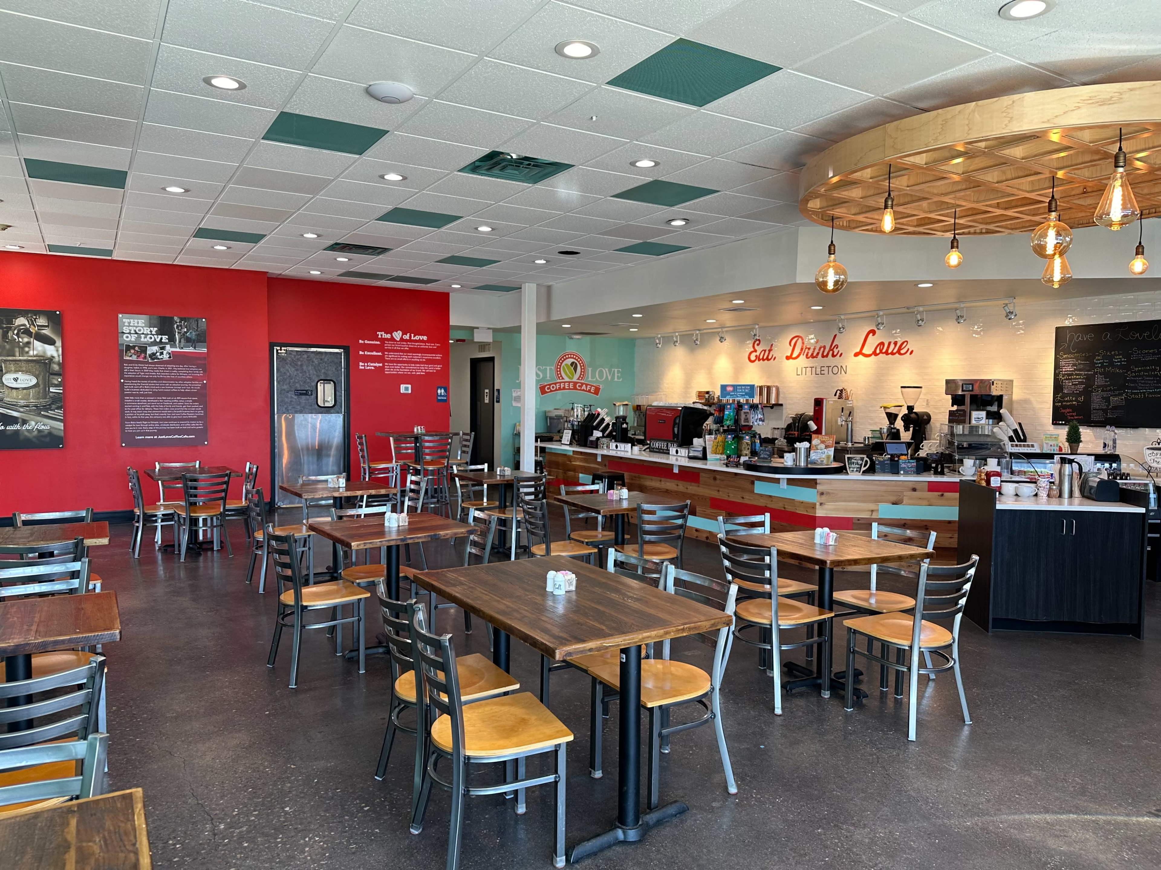 A modern café interior with wooden tables and metal chairs, a counter displaying a coffee station, and red-accented walls.