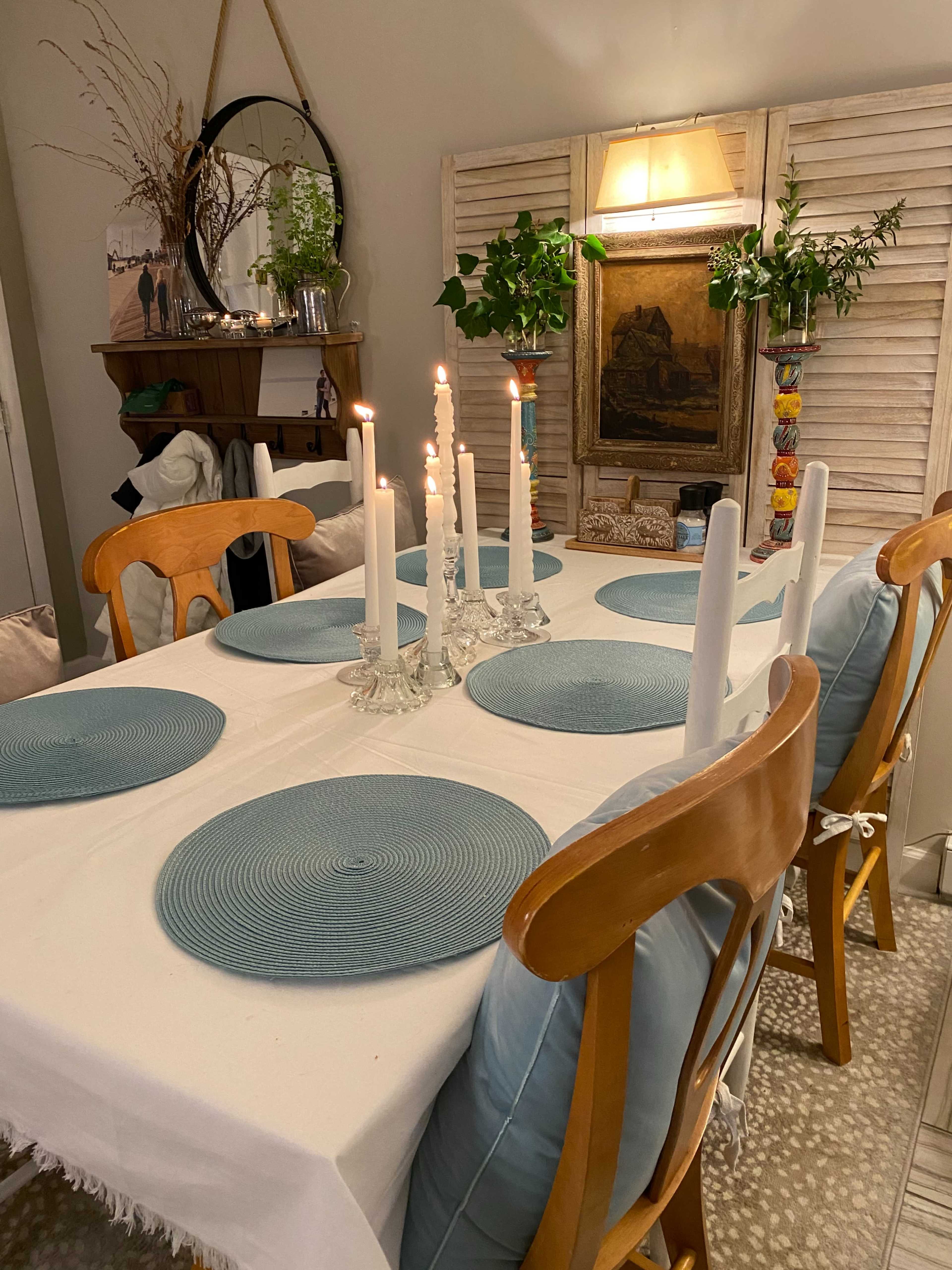 A dining table is set with blue placemats and white candles, surrounded by wooden chairs in a cozy, well-decorated room.