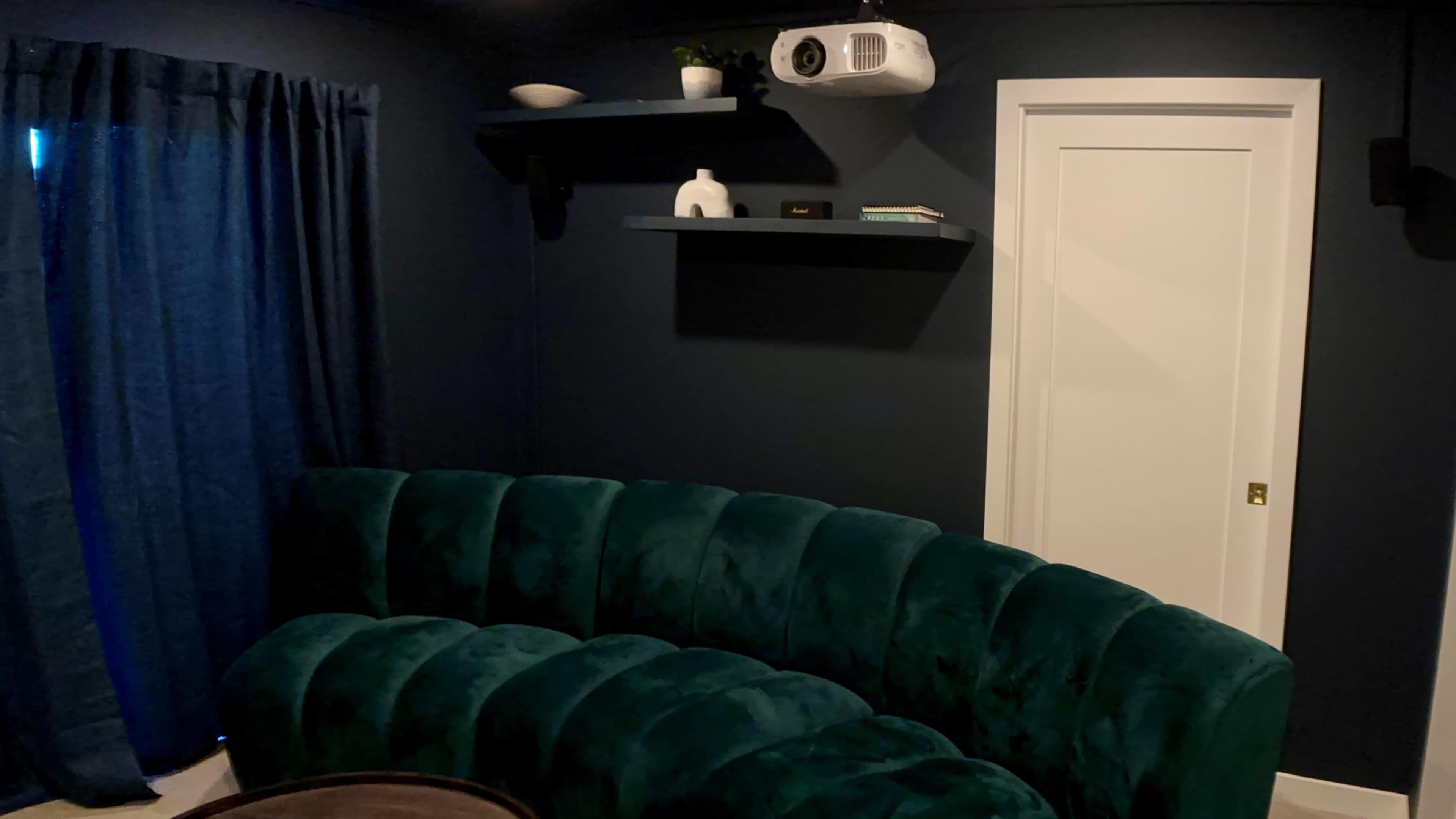 A curved green velvet sofa is positioned in a dark room with blue curtains, a wall-mounted projector, and a shelf displaying decorative items.