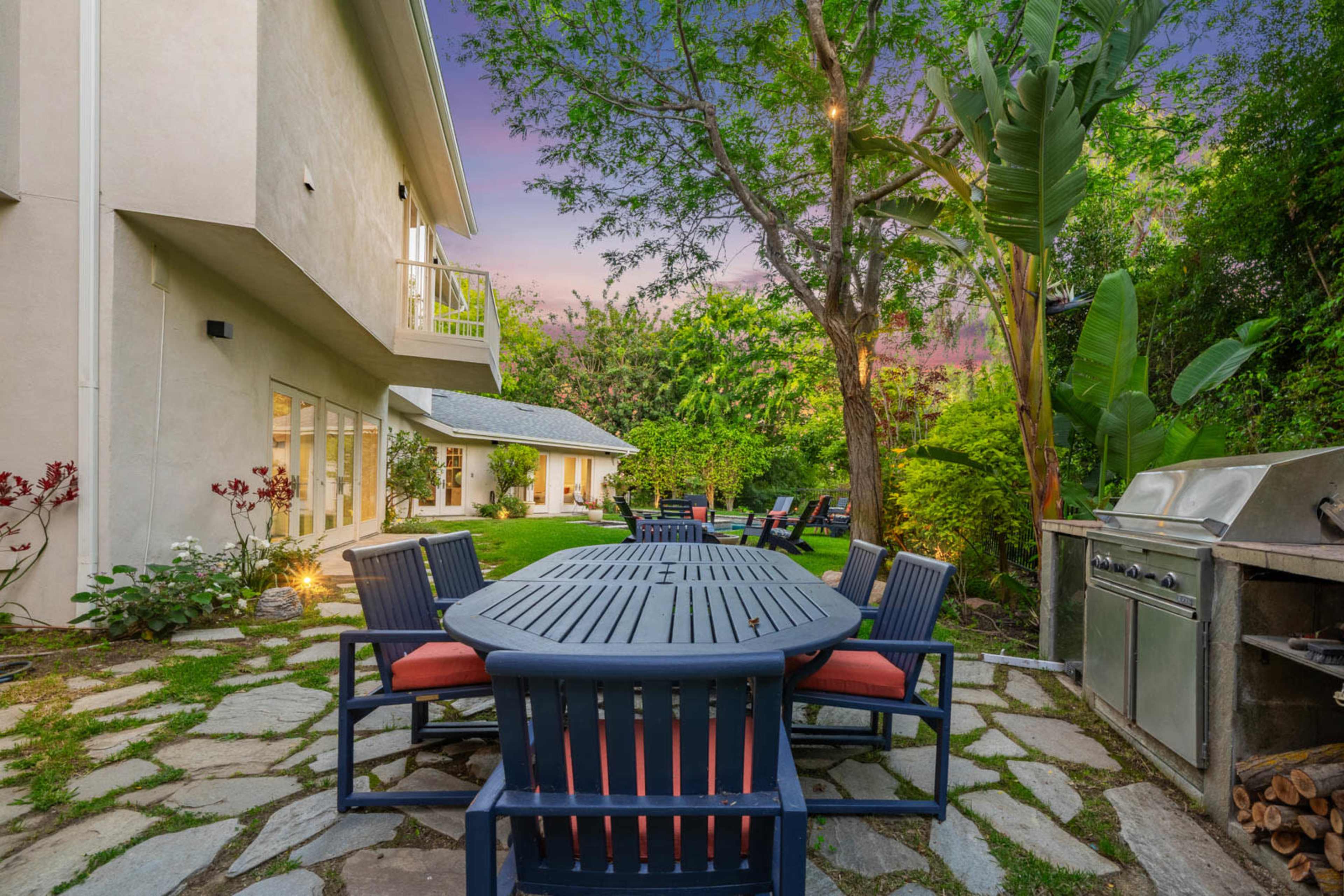 The image shows a patio area with a large dining table surrounded by blue chairs, a stone pathway, and a barbecue grill, set in a lush green garden.