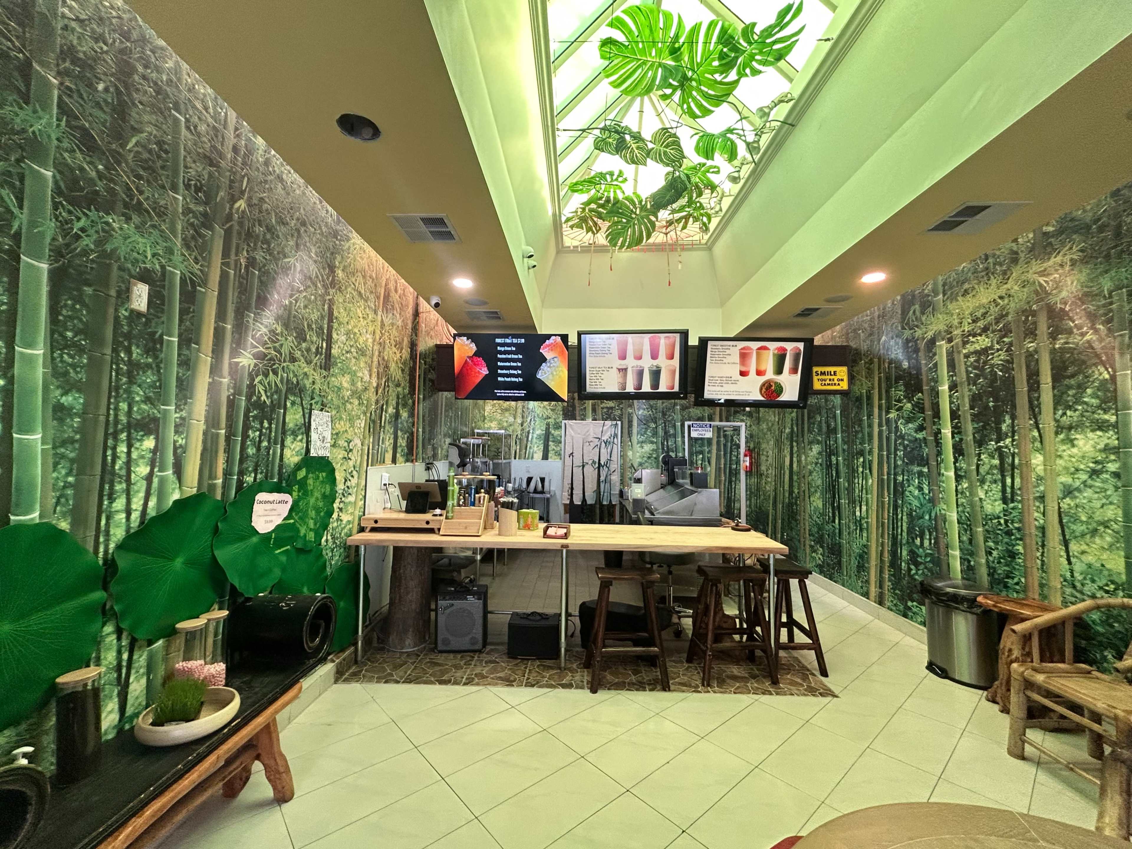 The image shows a café interior decorated with bamboo-themed wallpaper, featuring a counter with drink options displayed on screens and greenery overhead.