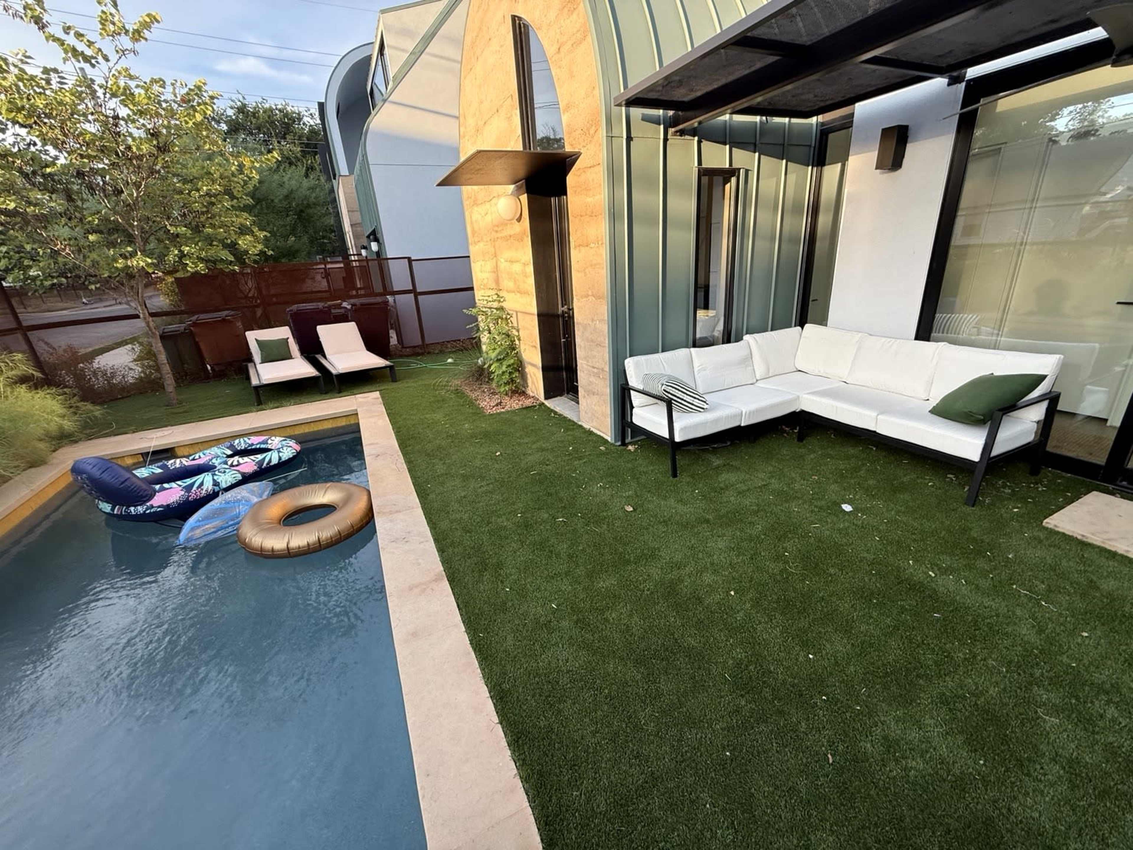 The image shows a backyard area featuring a small swimming pool, a white outdoor sofa set, two lounge chairs, and colorful inflatable floats on artificial grass.