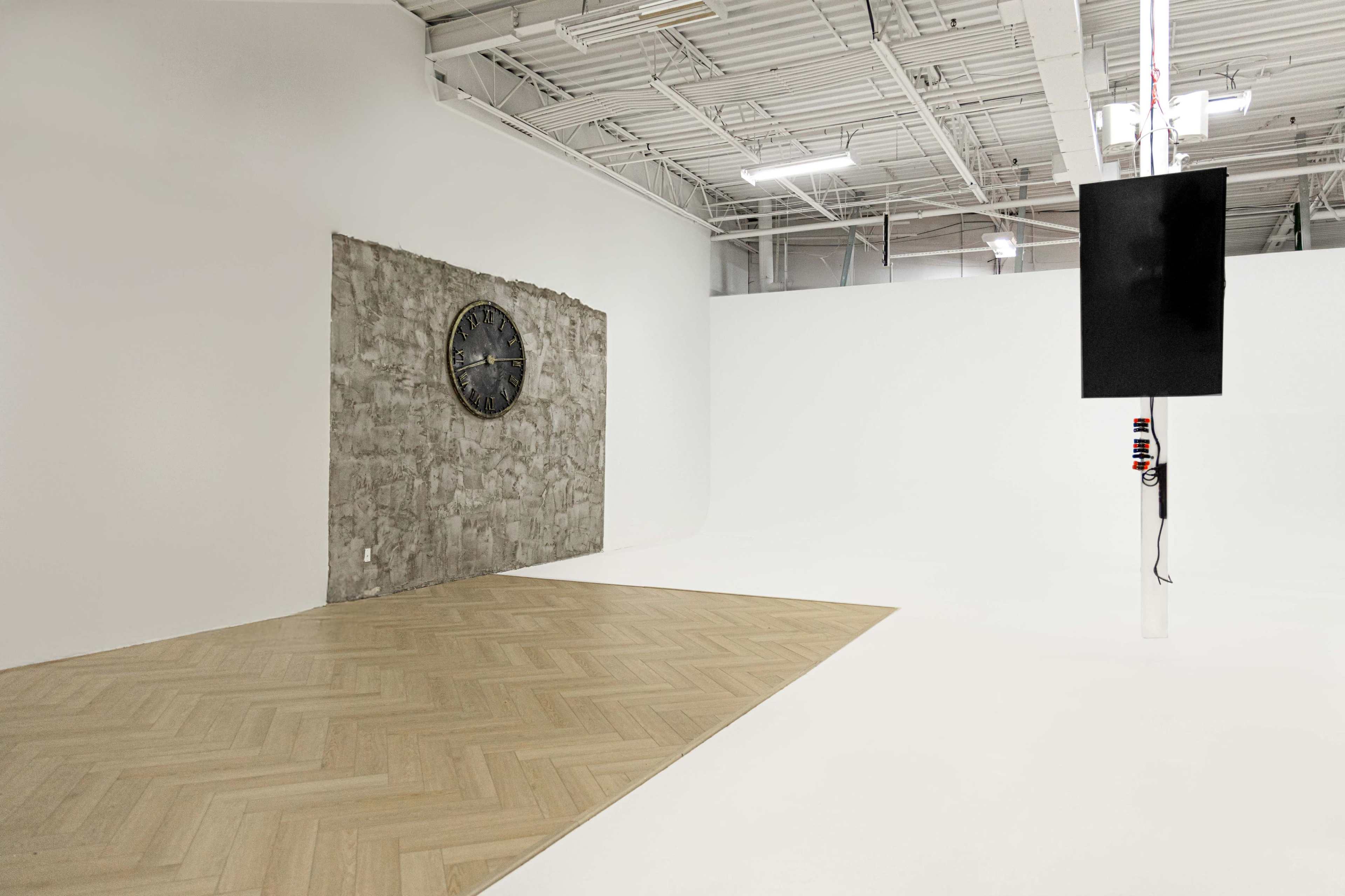 The image shows an empty interior space with a concrete wall featuring a clock, hardwood flooring, and a blank white background.