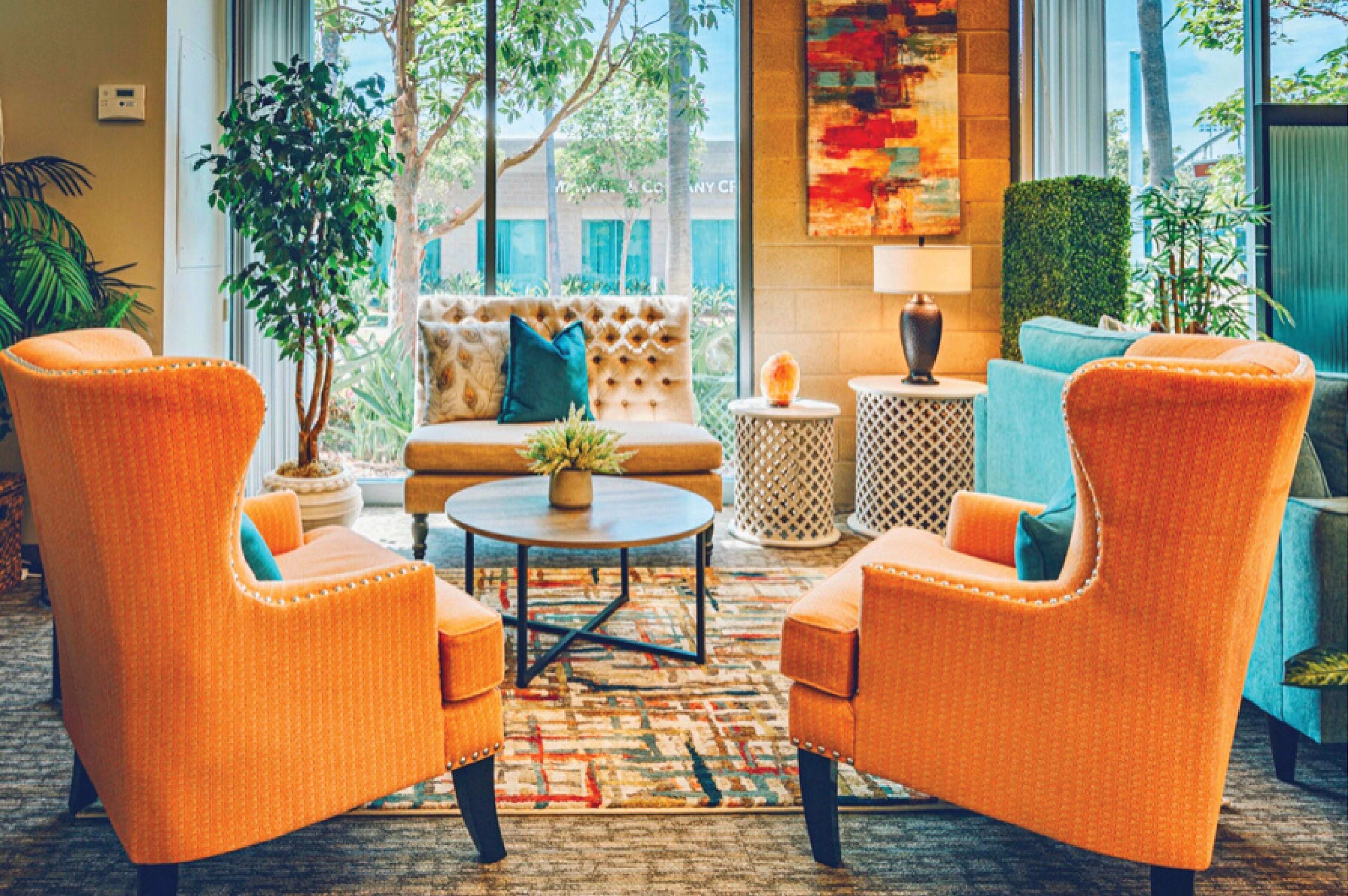 A bright and inviting lounge area featuring orange armchairs, a round coffee table, and a mix of greenery, with a colorful rug underfoot.