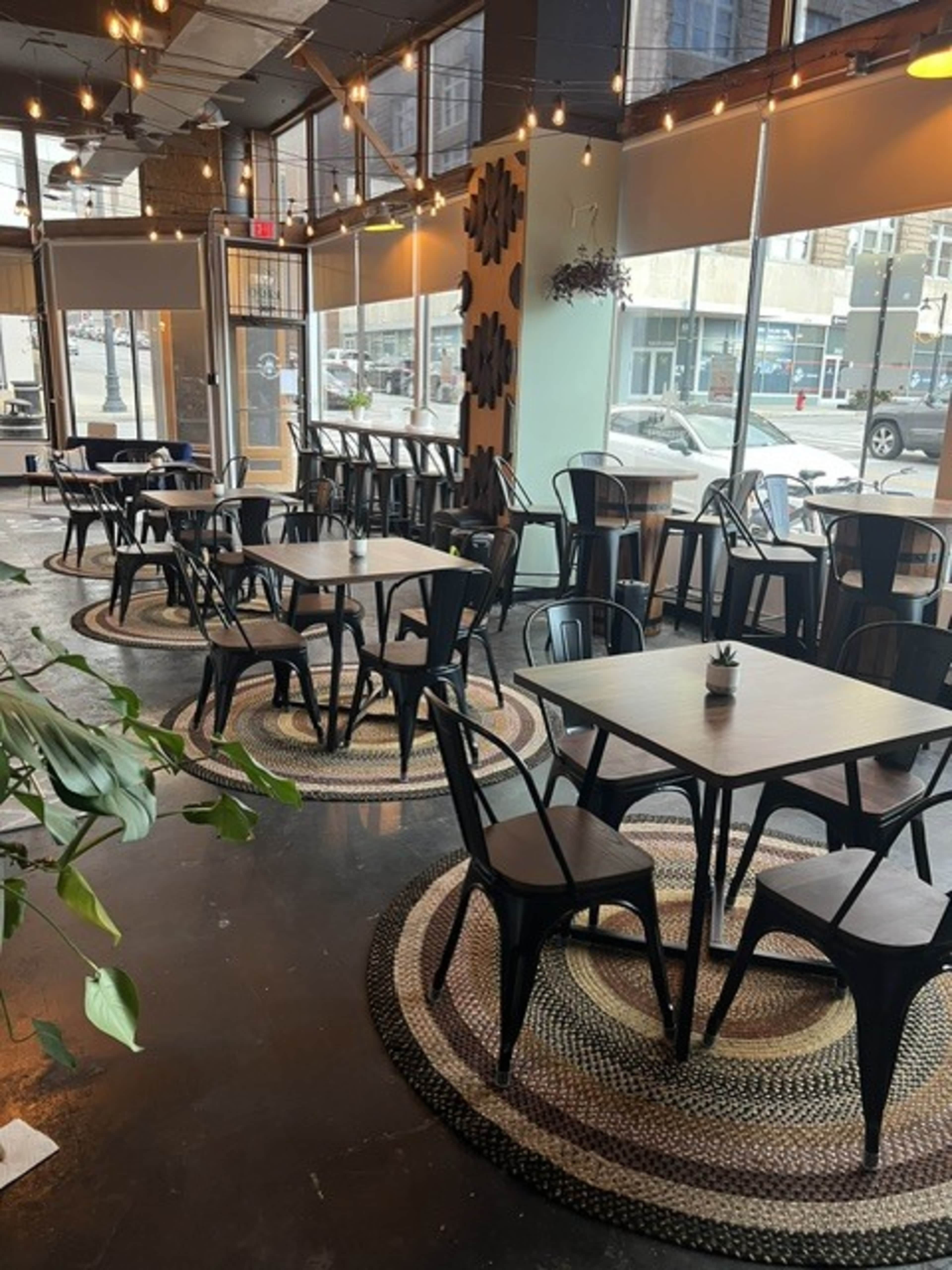 The image shows an interior of a café with black metal chairs and tables arranged on woven rugs, large windows letting in natural light, and modern decor elements.