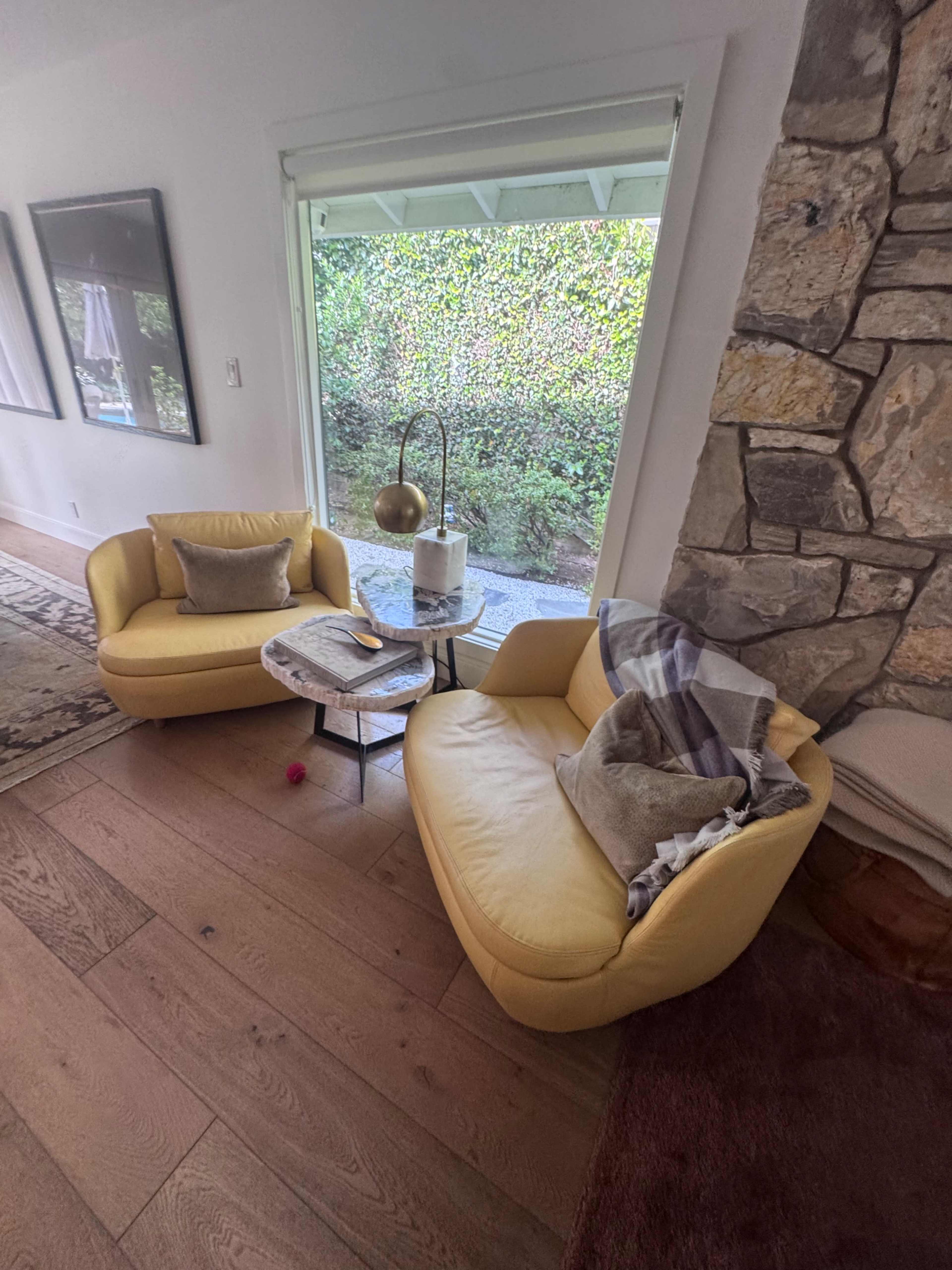 The scene features two yellow chairs positioned near a window, with a small table holding a book and a decorative lamp, set against a stone wall.